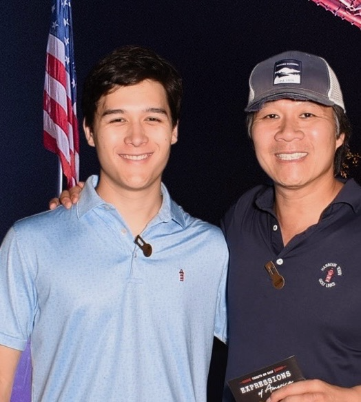 Liam Henry Pham, a young man in a light blue polo shirt with dark brown hair, standing with his father against a black backdrop with the American flag behind them