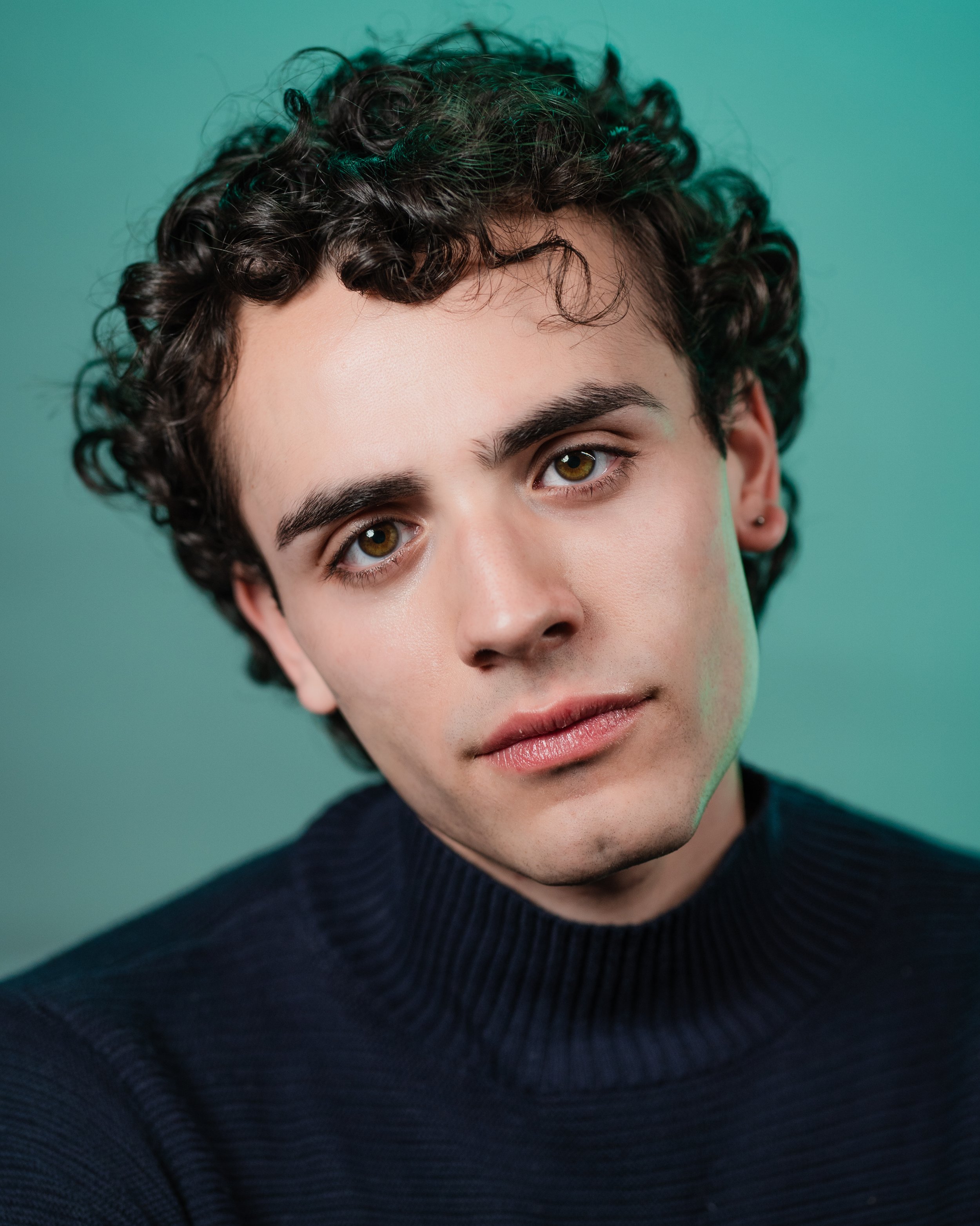 Jose-Emilio Escoto, a young actor with dark brown curly hair and brown eyes, in a navy sweater against a turquoise backdrop