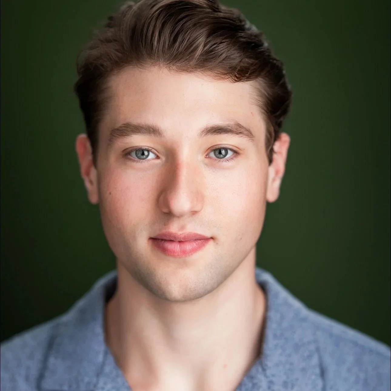 Close-up portrait of Jack Plogger, a young actor with blue eyes, brown hair, fair skin, wearing a gray shirt, against a dark green background.