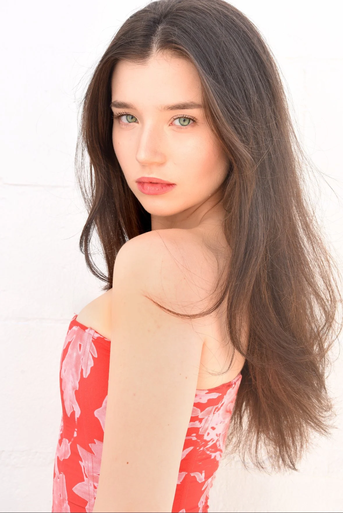 A young woman with long brown hair and green eyes looking over her shoulder, wearing a red and pink floral off-shoulder dress, against a plain white background.