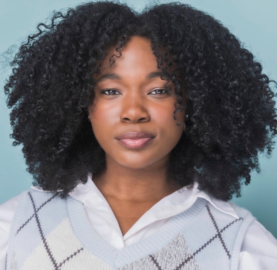 Rocky Anicette, a young actress with curly black hair, wearing a white collared shirt and a light gray argyle sweater, looking directly at the camera with a neutral expression against a light blue background.