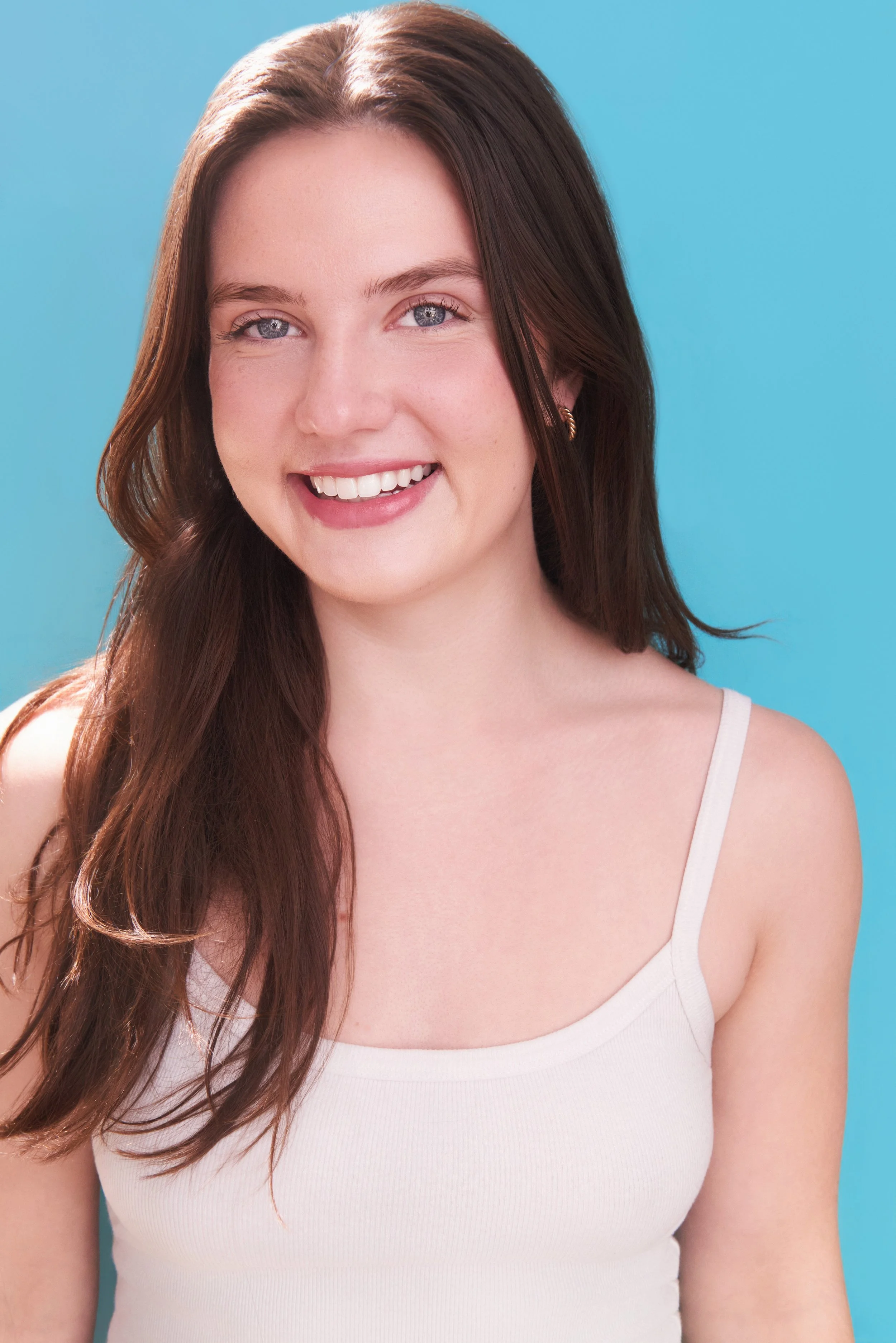 Elena Schlax, a young actress with brown hair and blue eyes, wearing a white tank top against a bright blue backdrop