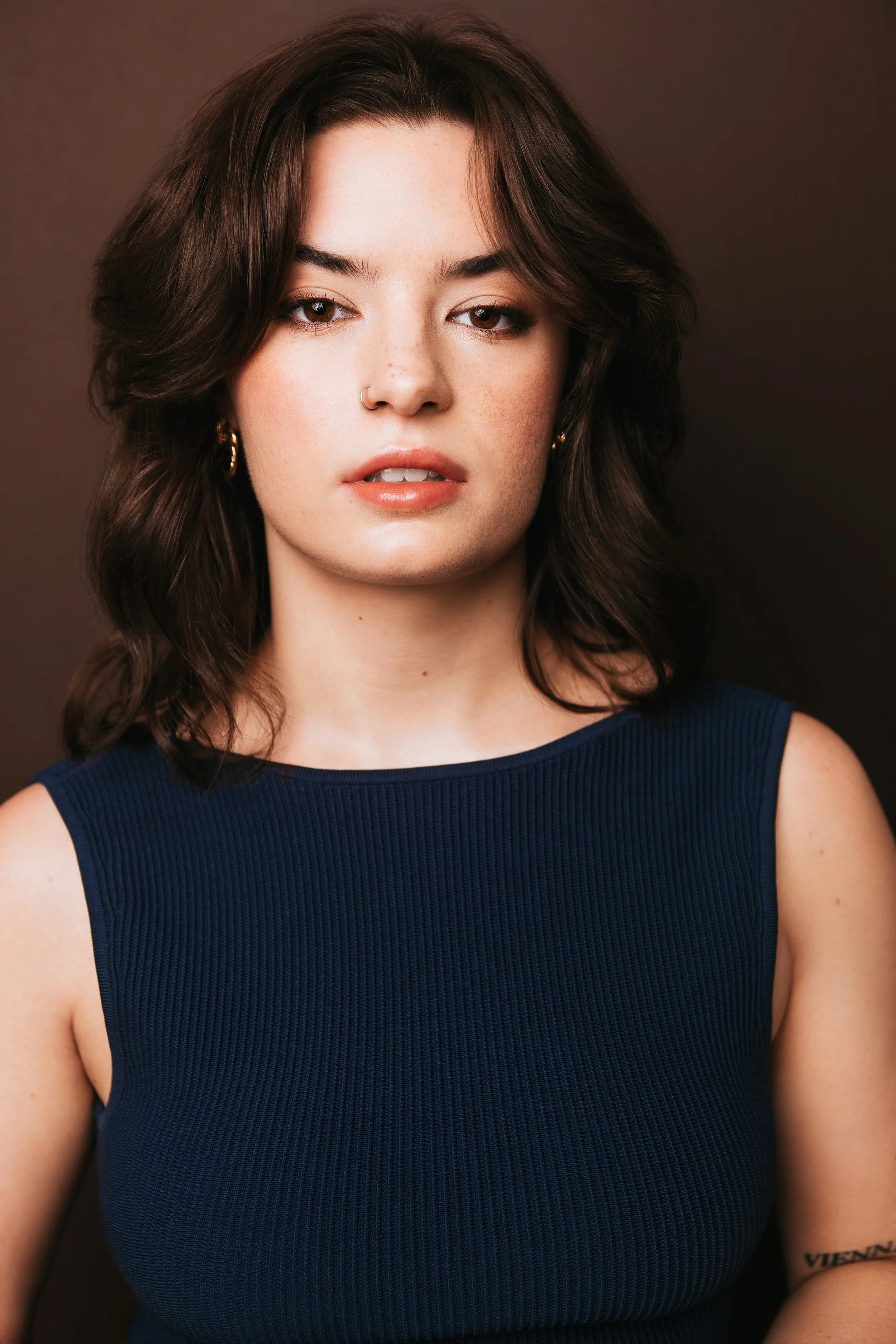 Close-up portrait of Kelsey LeVie, a young actress with short, wavy dark brown hair, wearing a navy blue sleeveless top, gold hoop earrings, and a nose ring against a dark background.