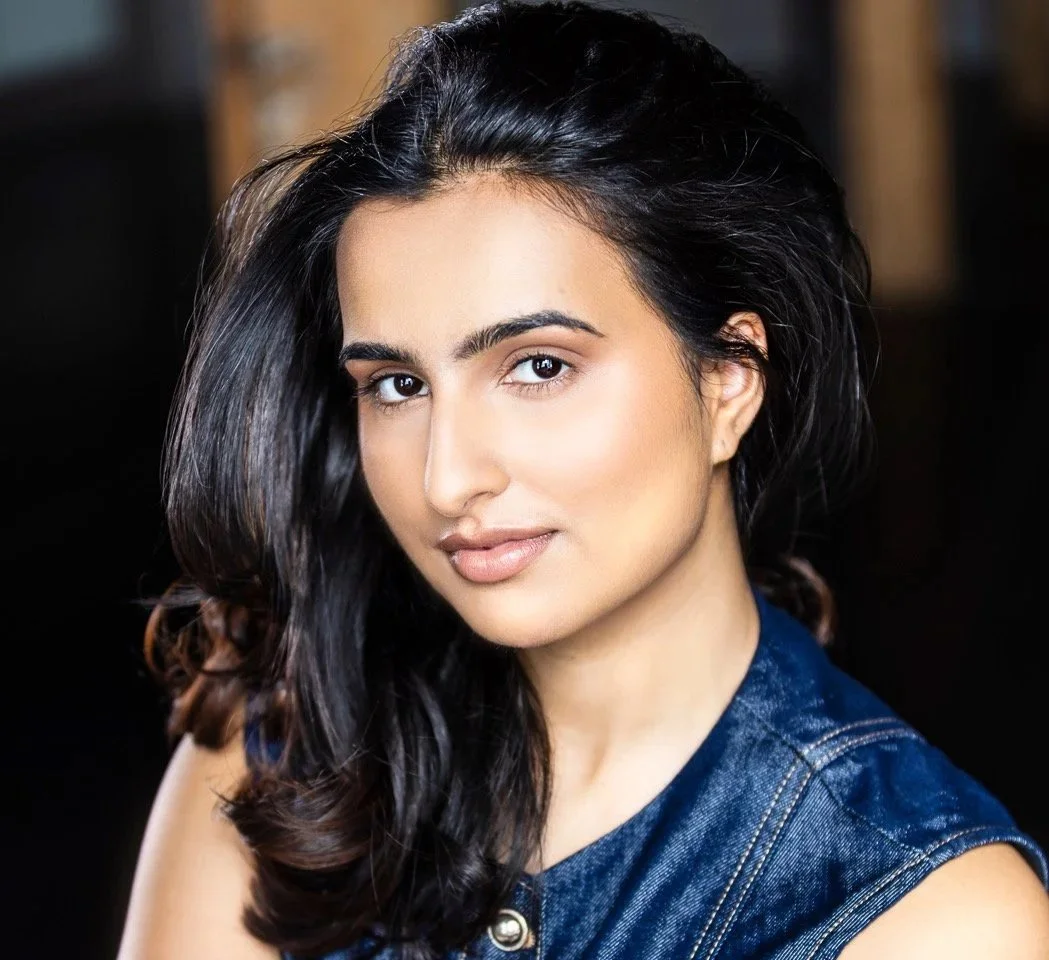 Uma Bains, a young actress with long, dark, wavy hair and light makeup, wearing a sleeveless denim top, looking at the camera with a slight smile.
