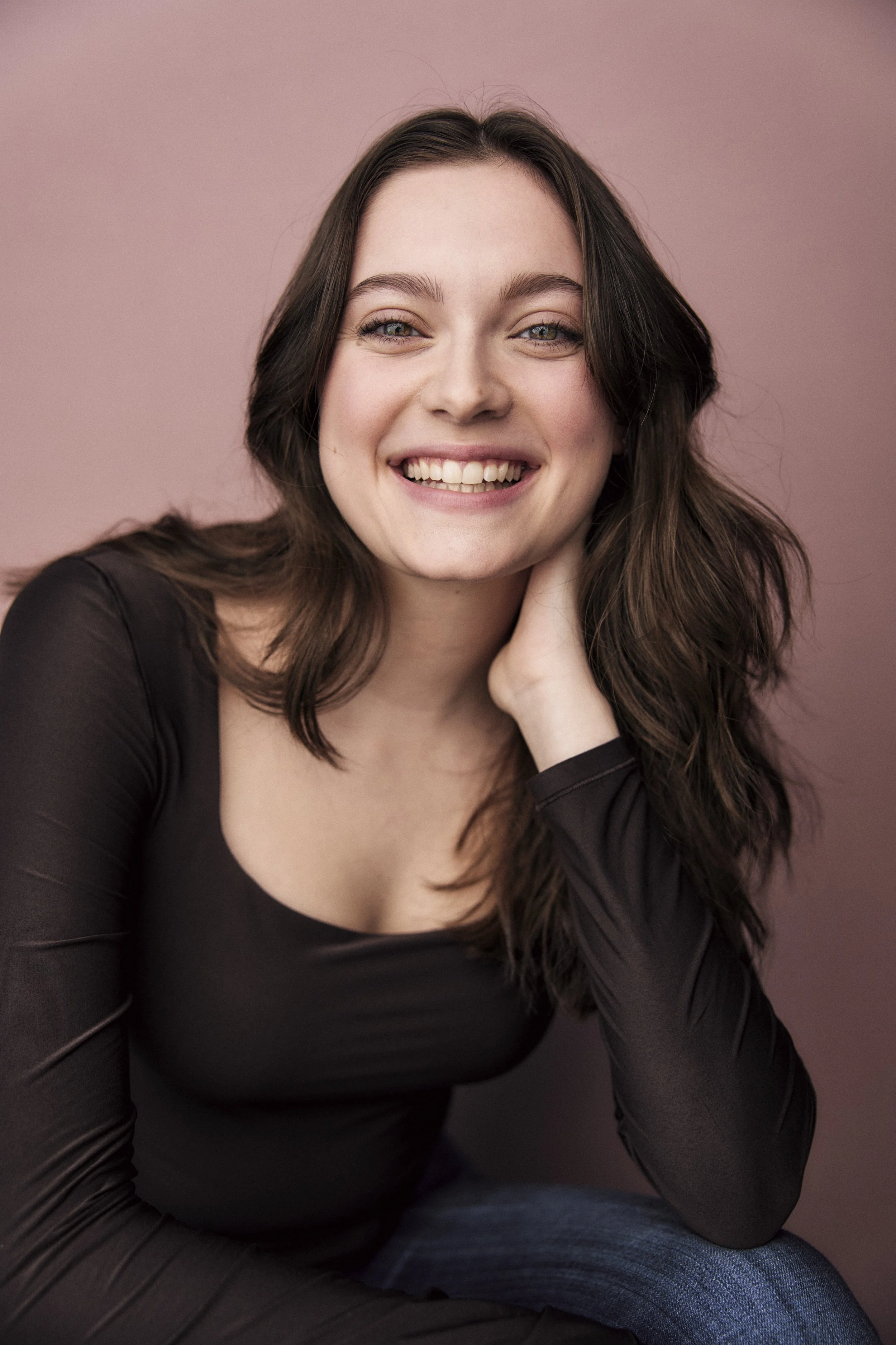Carolyn Dunbar, a young actress with wavy brown hair and brown eyes, smiling and leaning on her hand in a dark brown top against a pink backdrop