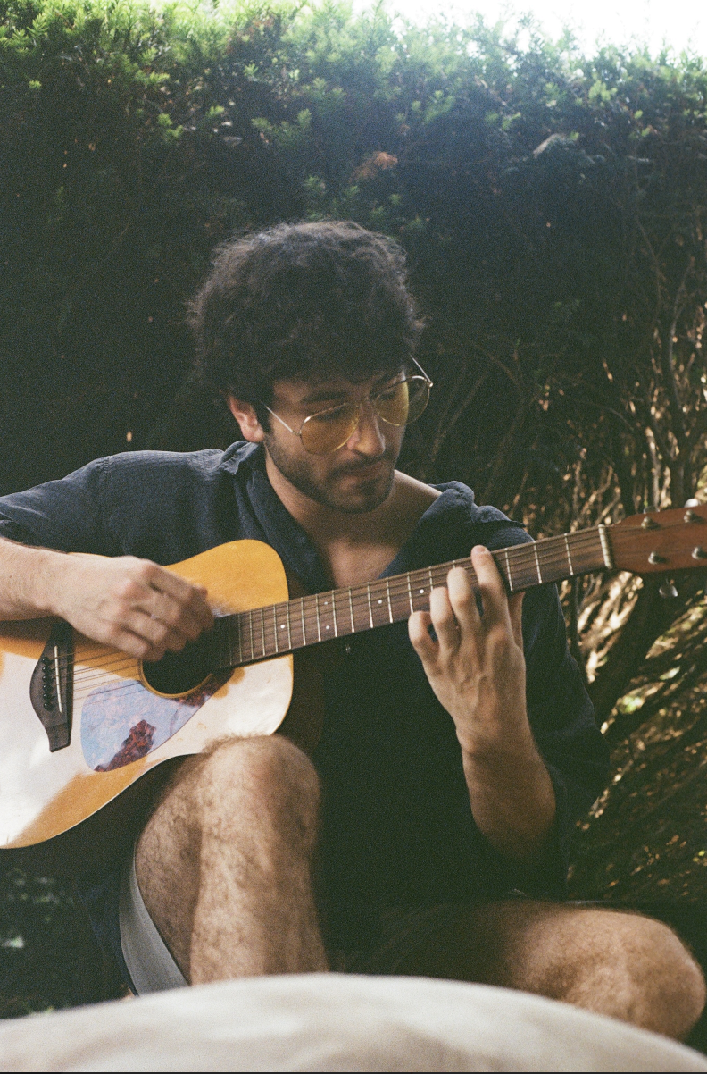 JJ Razzano, a young man with dark curly hair and yellow-tinted glasses playing an acoustic guitar outdoors, surrounded by green foliage.