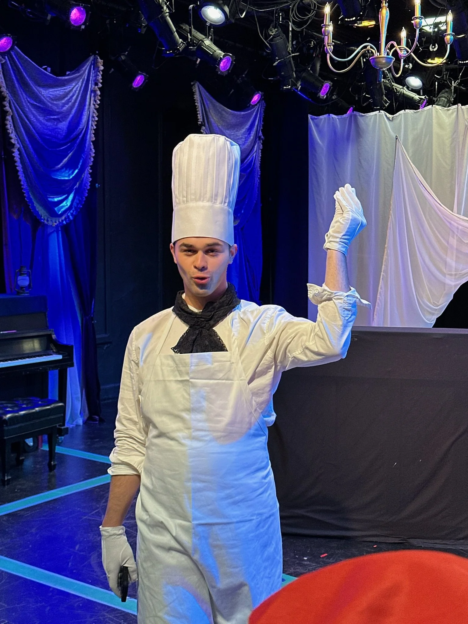Nathan Gutman, a young actor, dressed in a chef costume with a tall chef hat, white coat, and black necktie, standing on stage with a dark background and purple stage lights, holding a knife with their right hand raised.