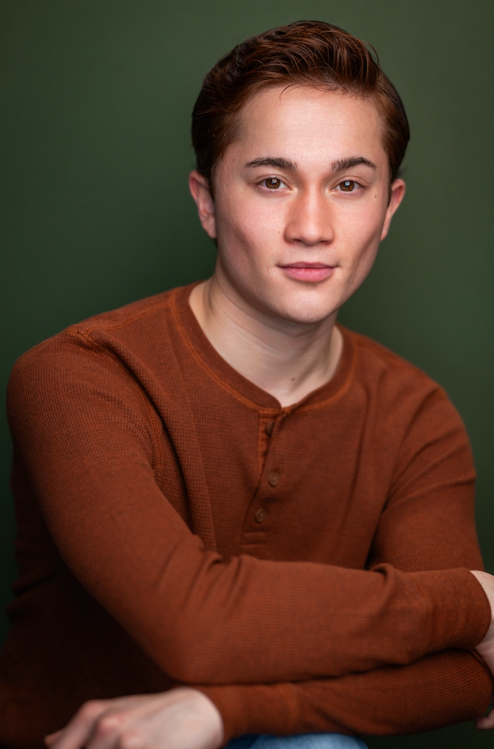Noah Ong Bamola, a young actor with ginger hair and brown eyes, poses calmly against a green backdrop with an orange shirt on