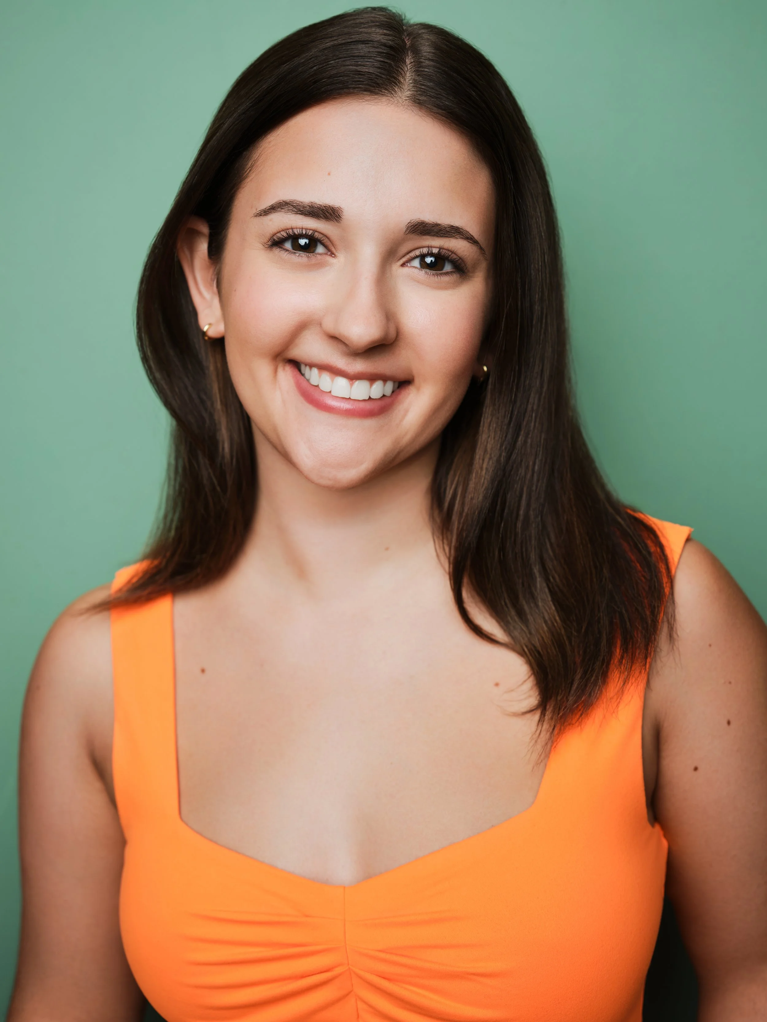 Jessica Schuchardt, a young actress with brown hair and dark brown eyes, smiles in a bright orange top against a turquoise backdrop