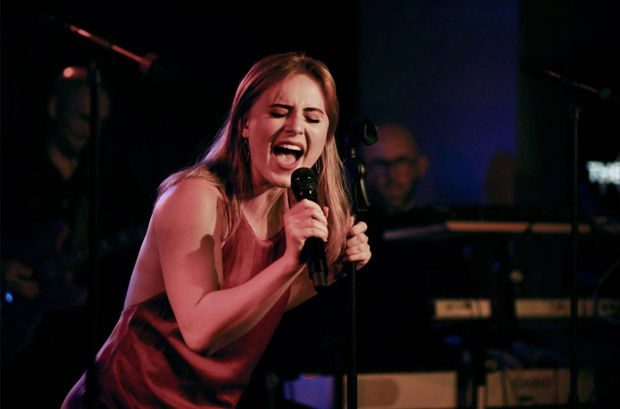 Isabella Victorson, a young actress and singer with blonde hair, singing with a microphone during a live performance and wearing a red satin dress