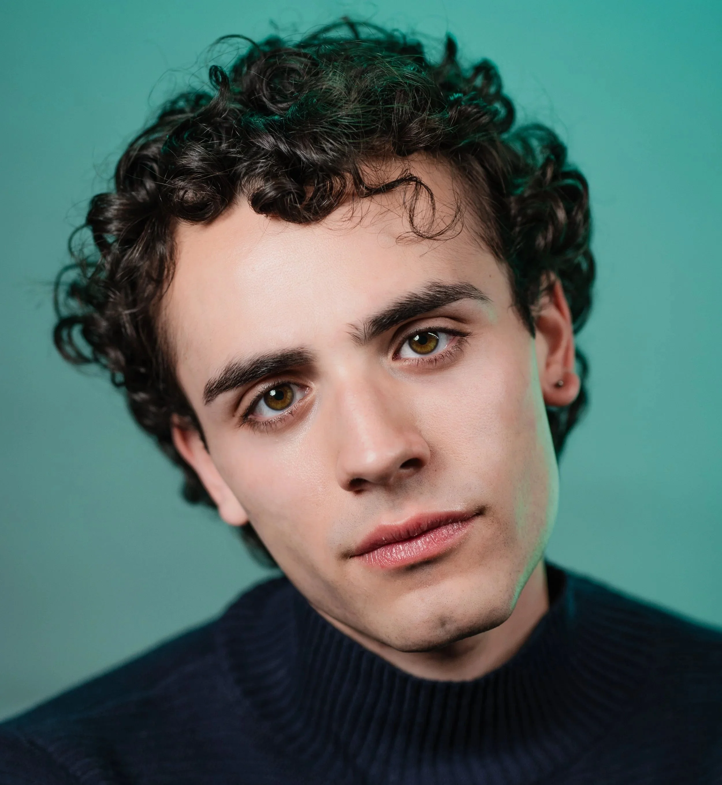 Close-up of Jose-Emilio Escoto a young actor with short, curly black hair, olive skin, and hazel eyes, wearing a black sweater, looking directly at the camera against a teal background.