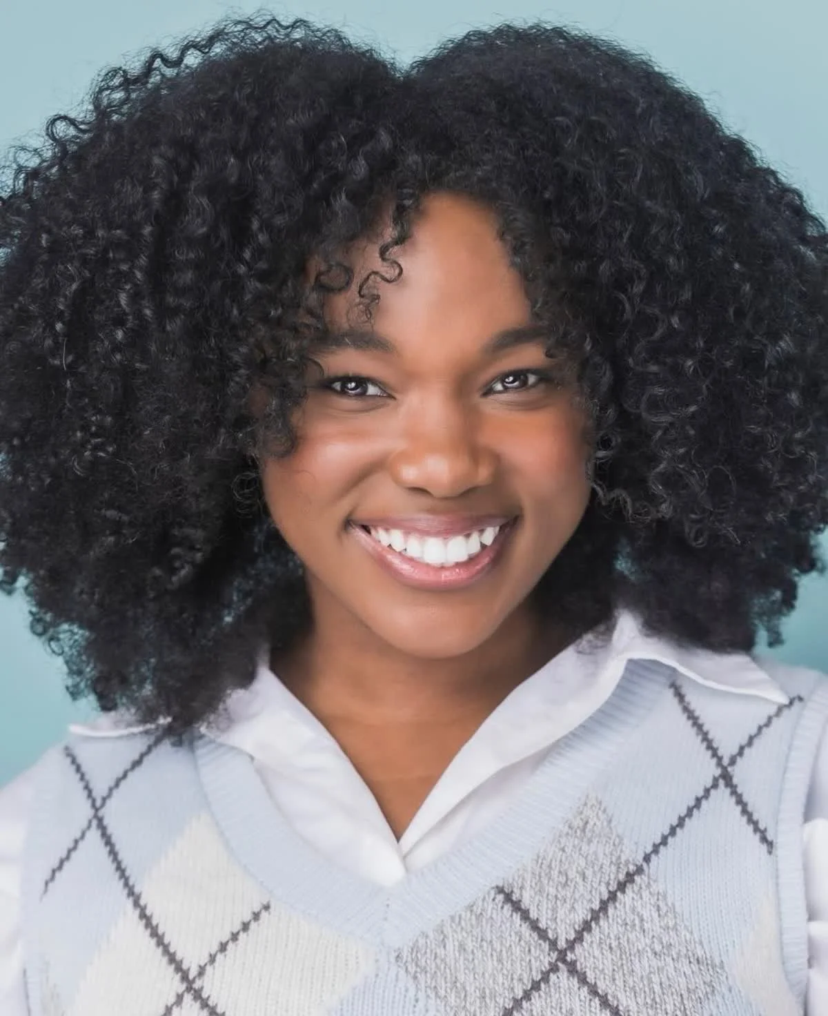 Close-up portrait of Rocky Anicette, a smiling young actress with curly hair wearing a light gray sweater with a diamond pattern and a collared shirt underneath against a light blue background.