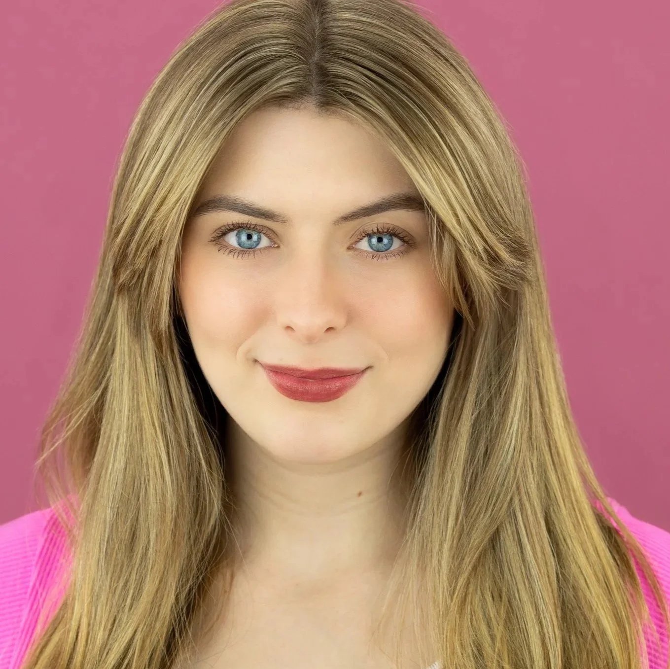 Close-up of Ruby Griffin, a young actress with long blonde hair, blue eyes, and pink lipstick, wearing a pink top, against a pink background.