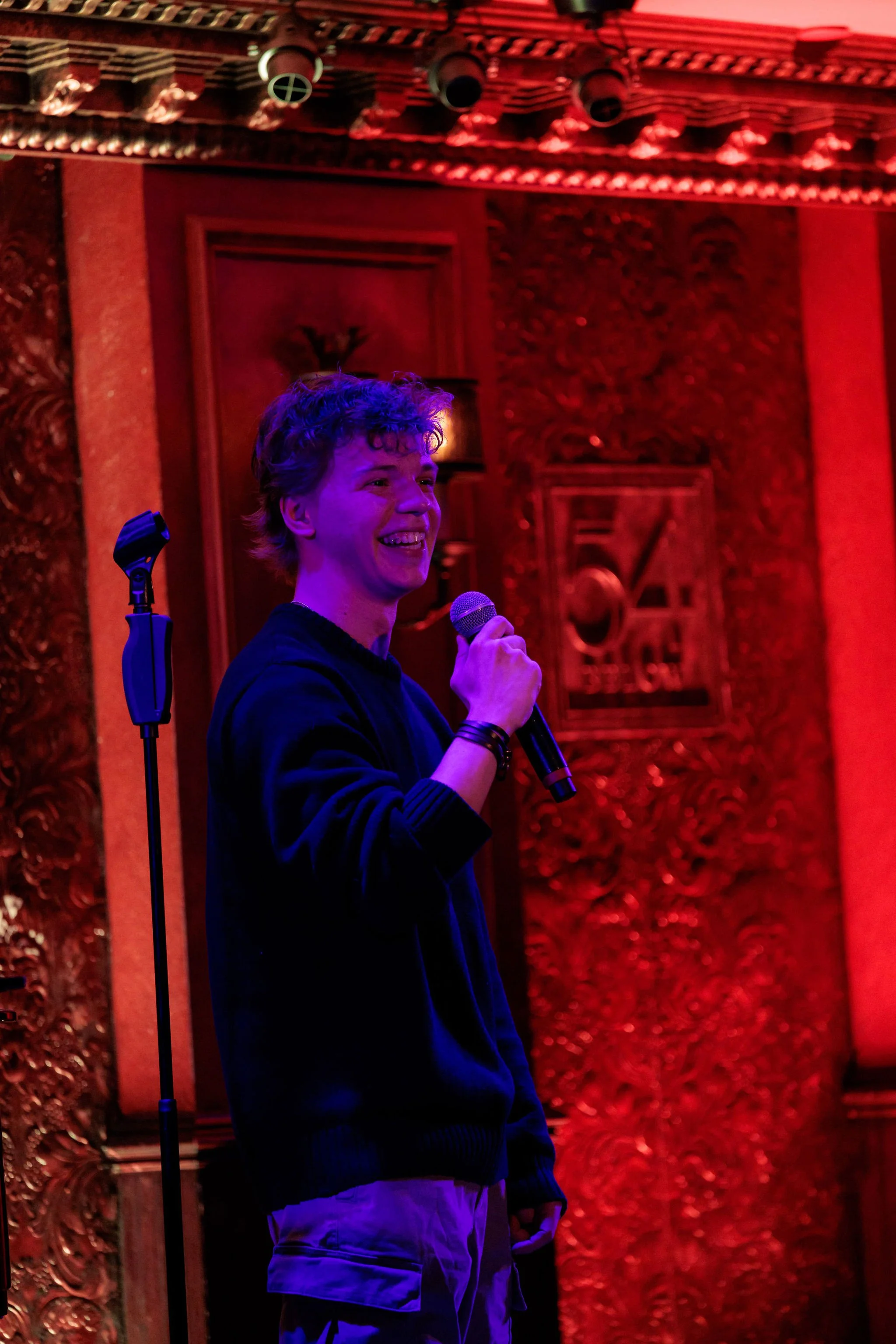 A young man with curly hair smiling and holding a microphone in a red-lit room with ornate woodwork in the background.