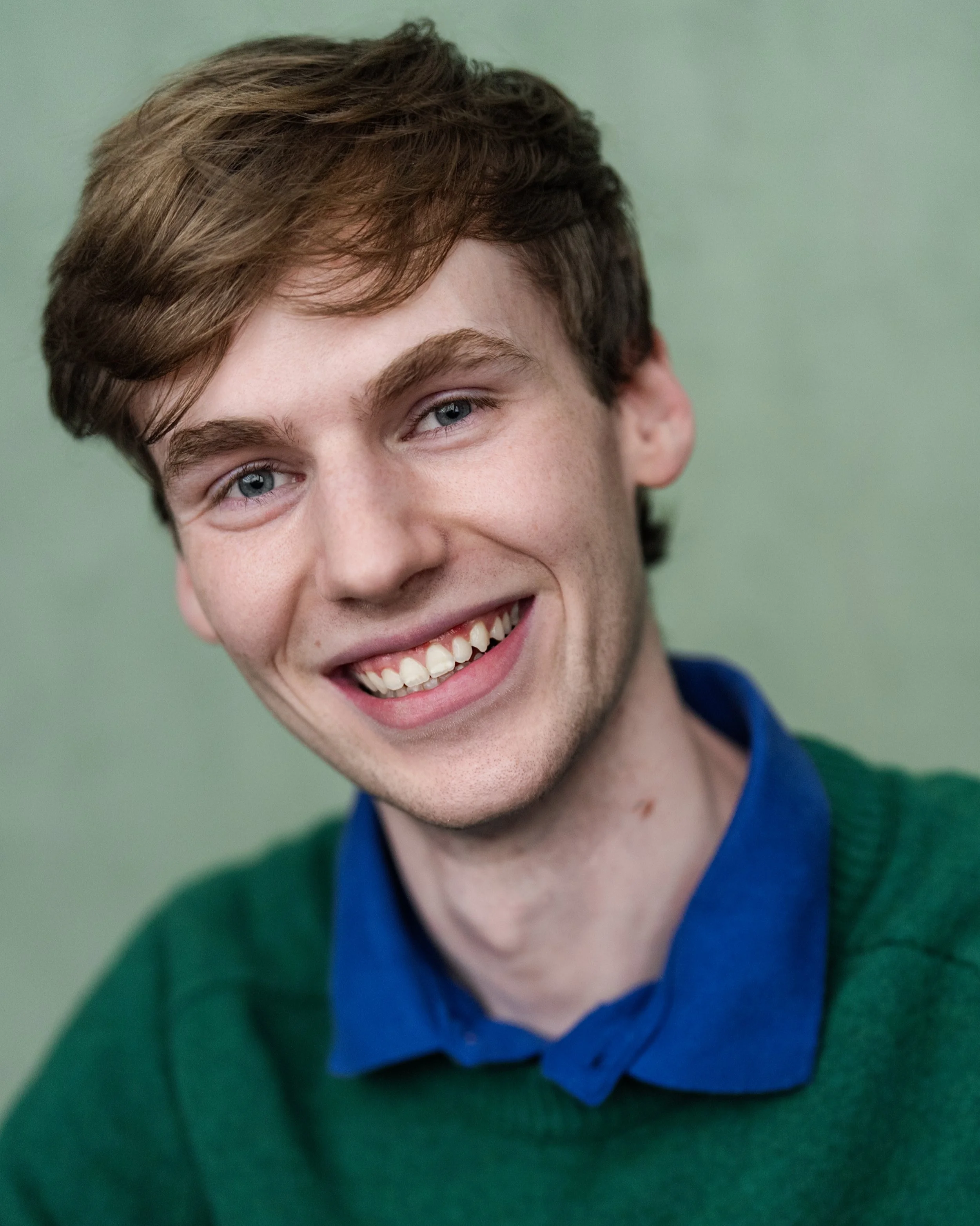 Aidan Ryan, an actor with blonde hair and blue eyes, in a green sweater with a blue collared shirt underneath, smiling against a light turquoise backdrop
