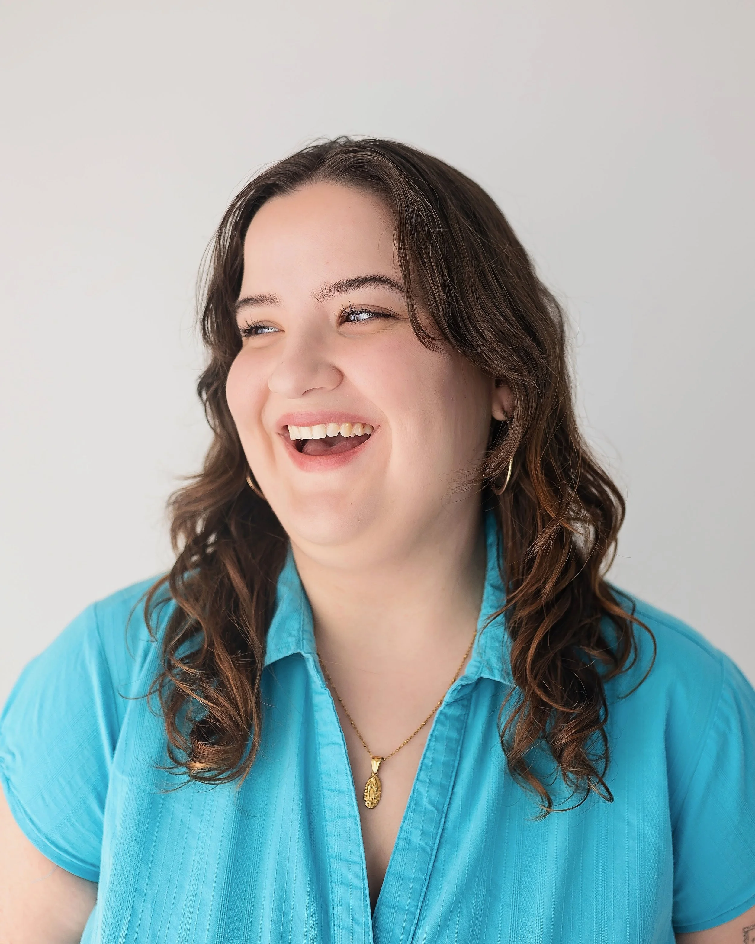 Gabriela Diaz, a young actress with wavy brown hair and brown eyes, wearing a bright blue top and gold necklace and laughing against a white backdrop