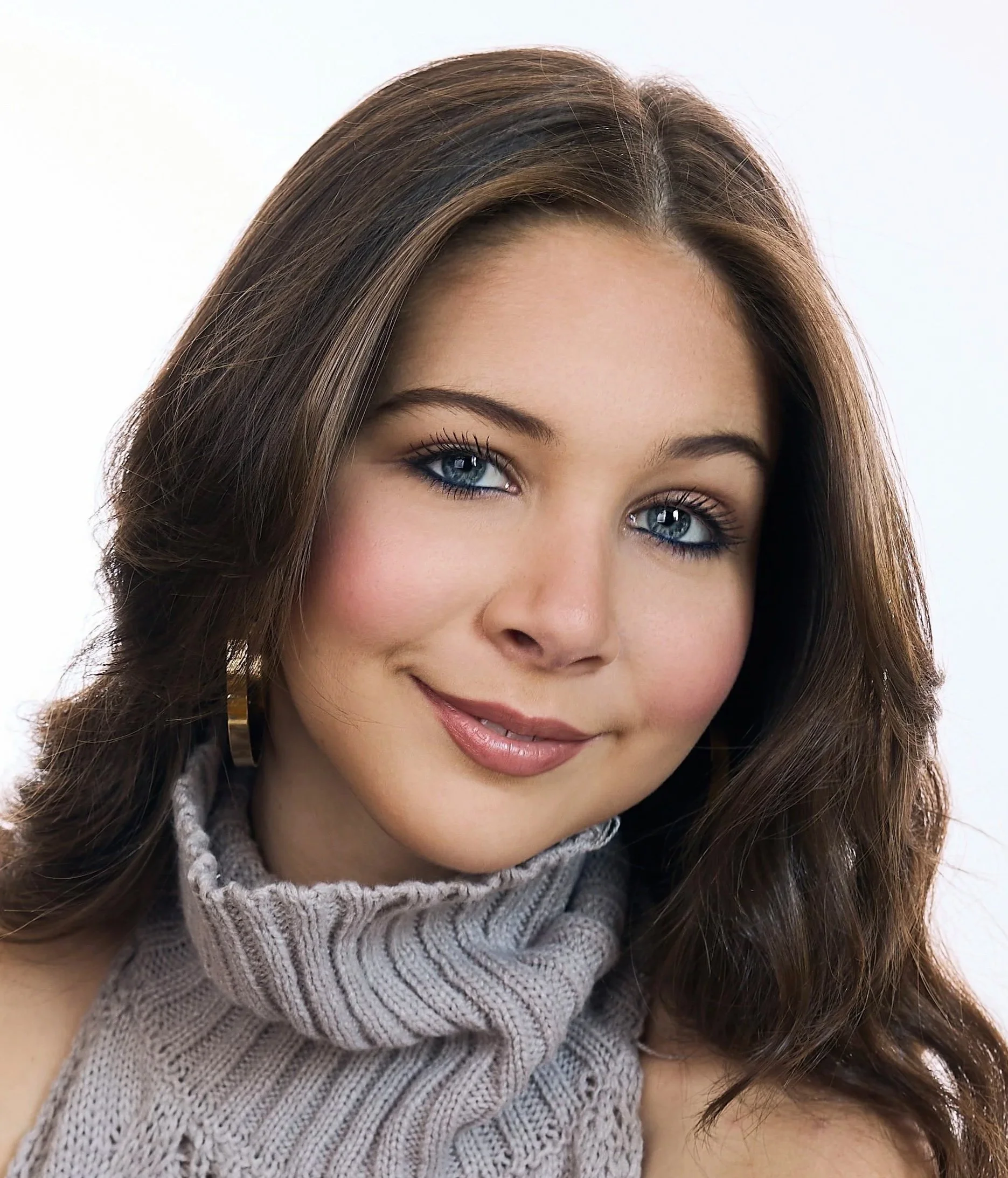 Close-up of Lucy McIver, a young actress, showing her blue eyes, brown hair with light highlights, and a smile, wearing a gray knitted sweater.