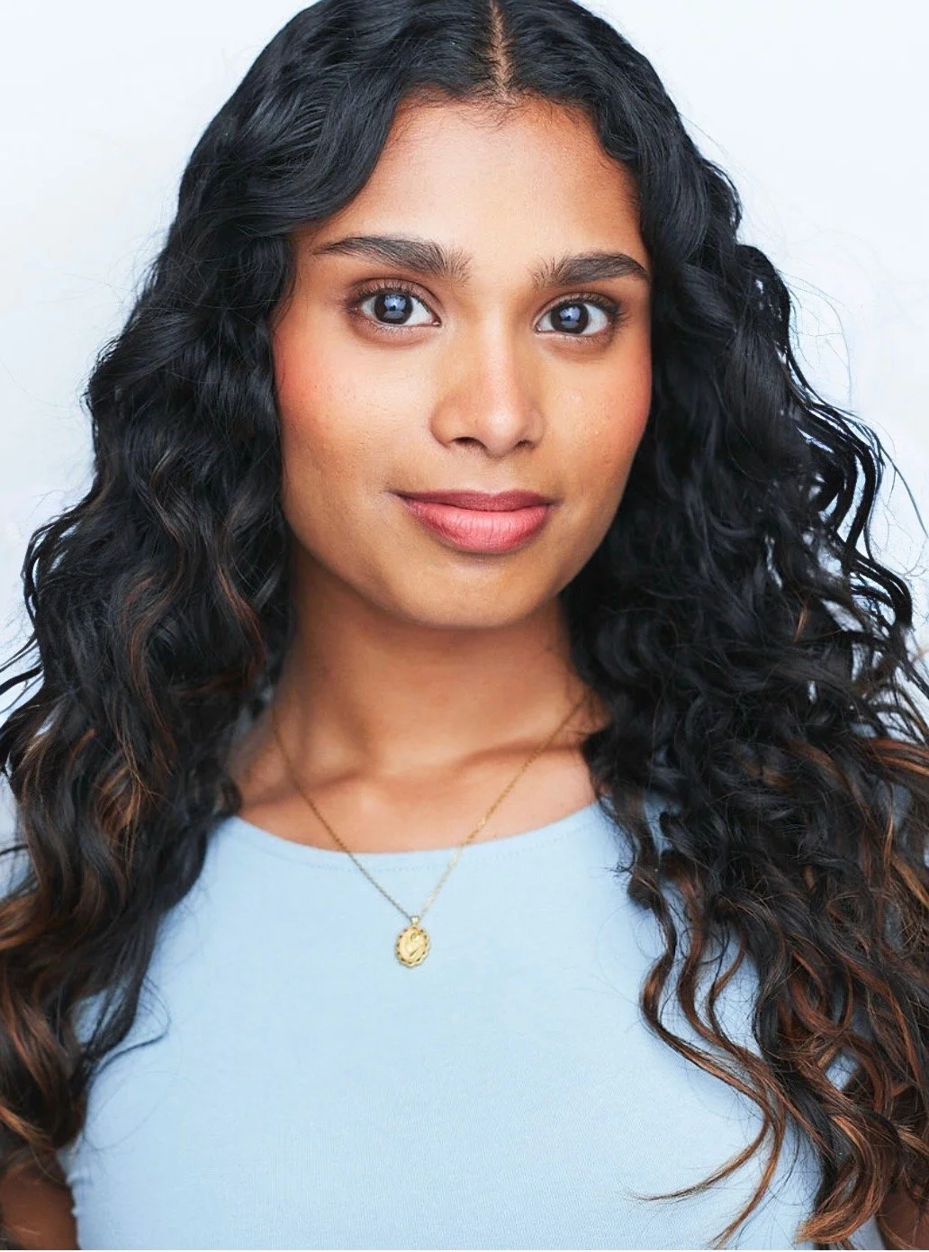 Becky Alex, a young South Asian actress with curly black hair and dark brown eyes in a light blue top and gold necklace against a white backdrop