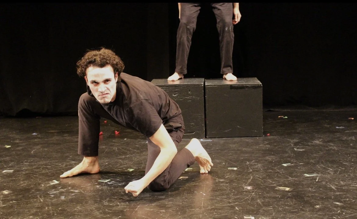 Jose-Emilio Escoto, a young actor with dark brown curly hair and brown eyes, crouched on the floor in an all black outfit during a performance of a self-written piece