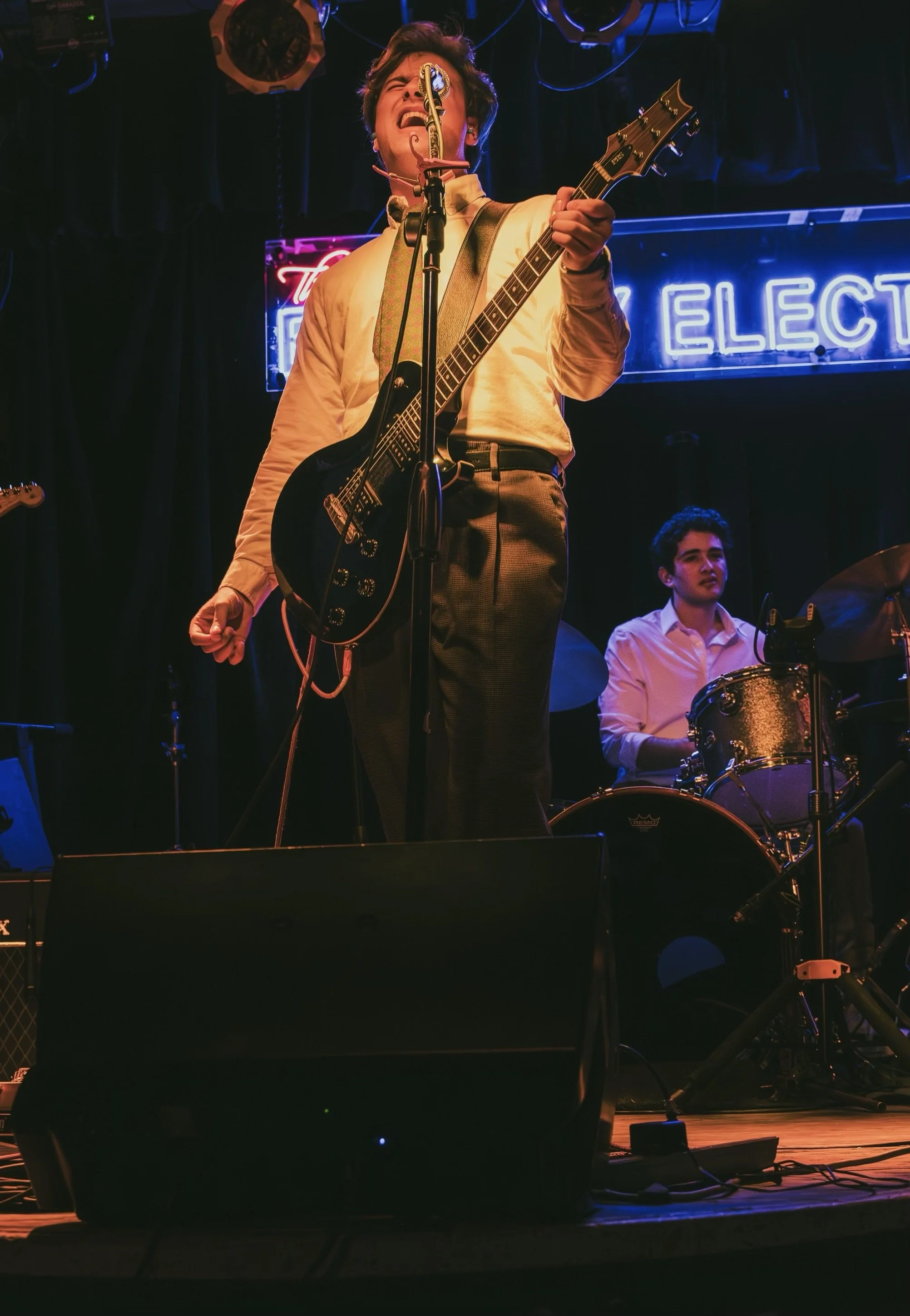 Matt Woodward, a young actor and musician with dark brown hair, standing on stage with a guitar and microphone in a white button down and dark jeans