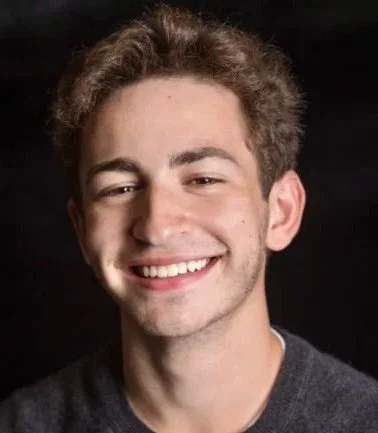 Davey Fried, a young actor with brown curly hair, smiling, against a dark background.