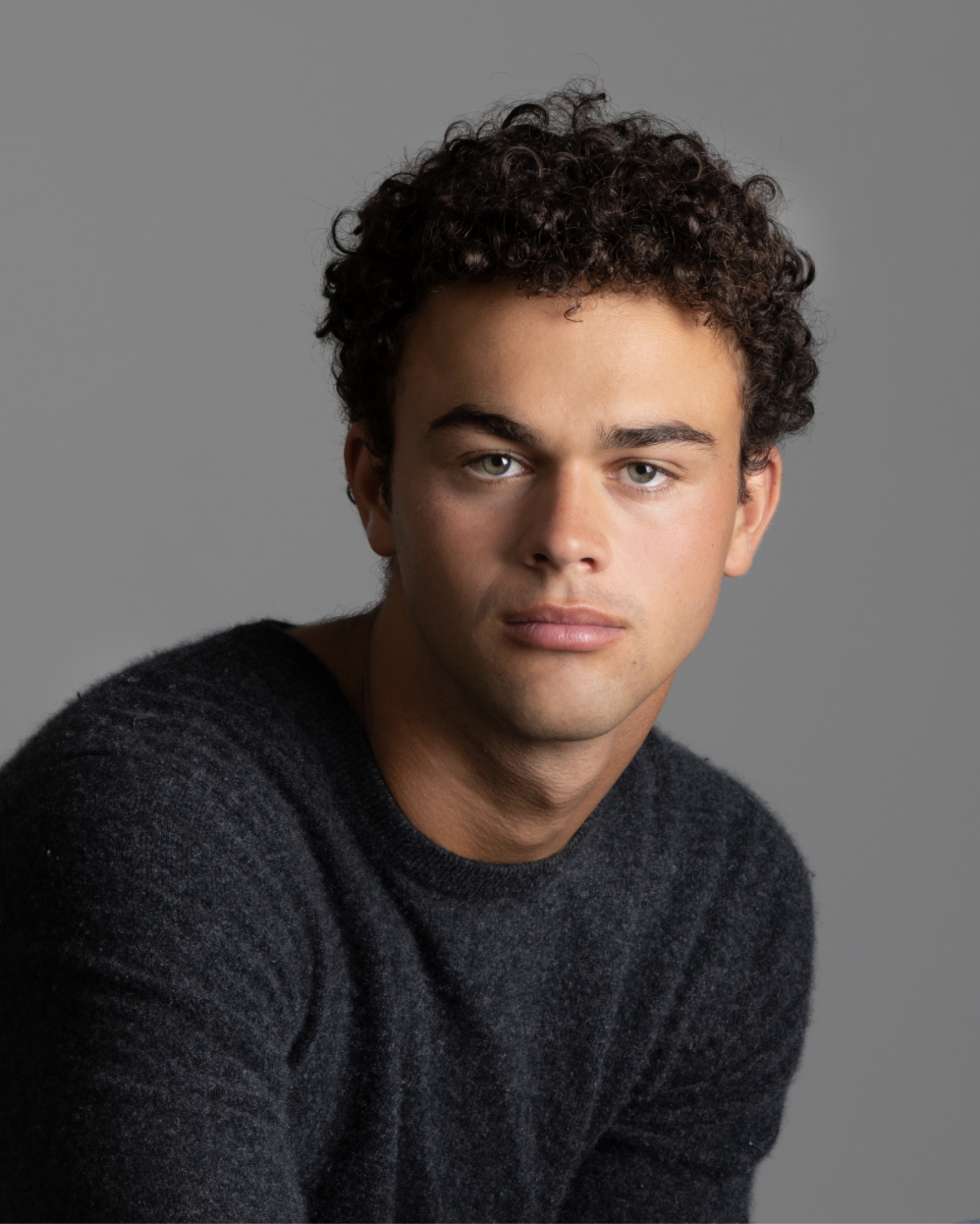 Nathan Gutman, a young actor with dark curly hair and light green eyes in a dark gray sweater against a gray backdrop