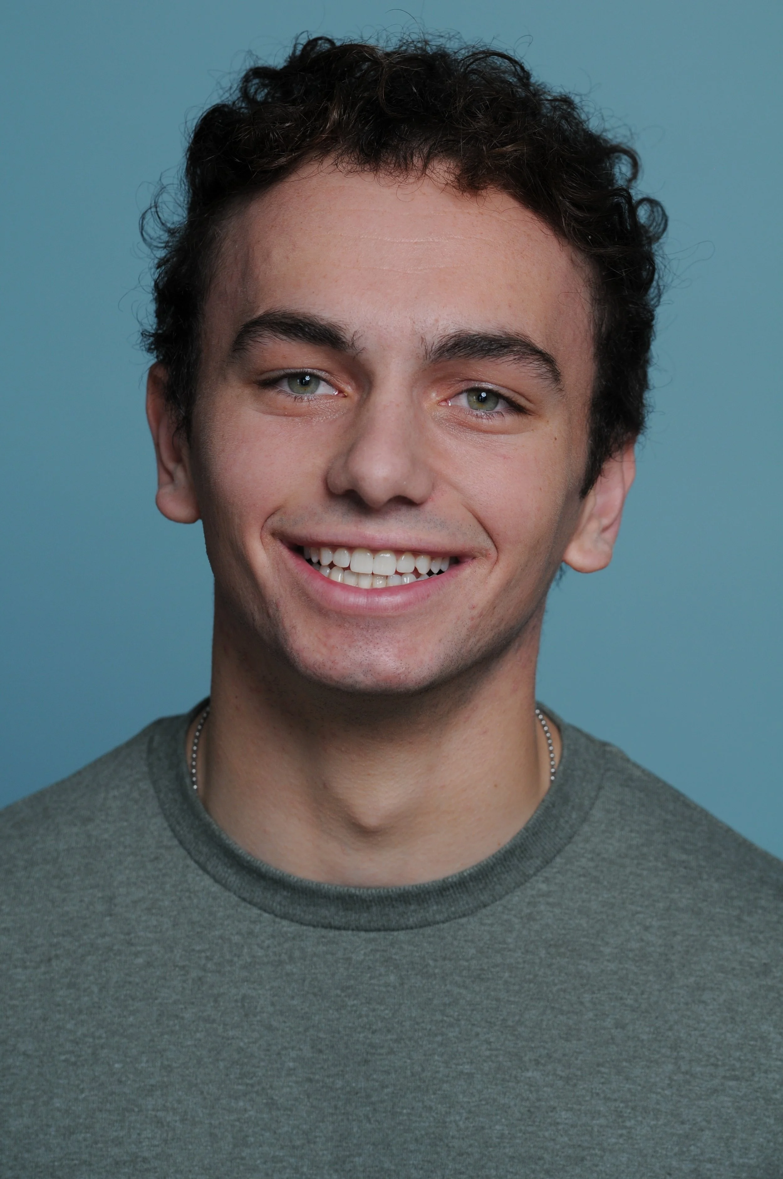 Liam Restivo, a young actor with curly brown hair and green eyes smiles in a gray tee shirt against a light blue backdrop