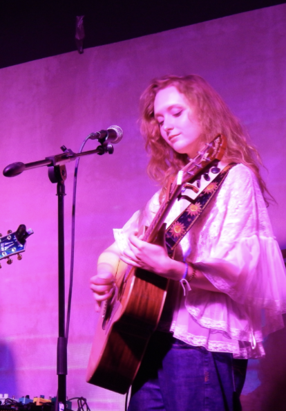 Grace Church, a young actress and musician with wavy ginger hair playing a guitar into a microphone wearing a flowy white lace top