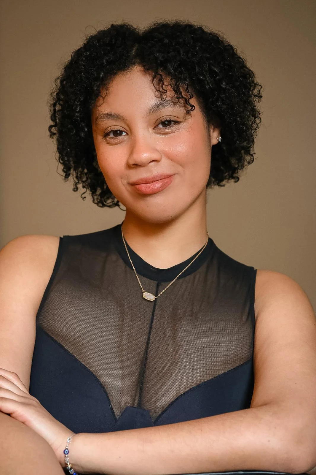 Sawyer Evans, a young actress with curly black hair smiling, wearing a black sleeveless top with sheer fabric, a gold necklace with a pendant, and a matching bracelet, standing against a plain beige background.