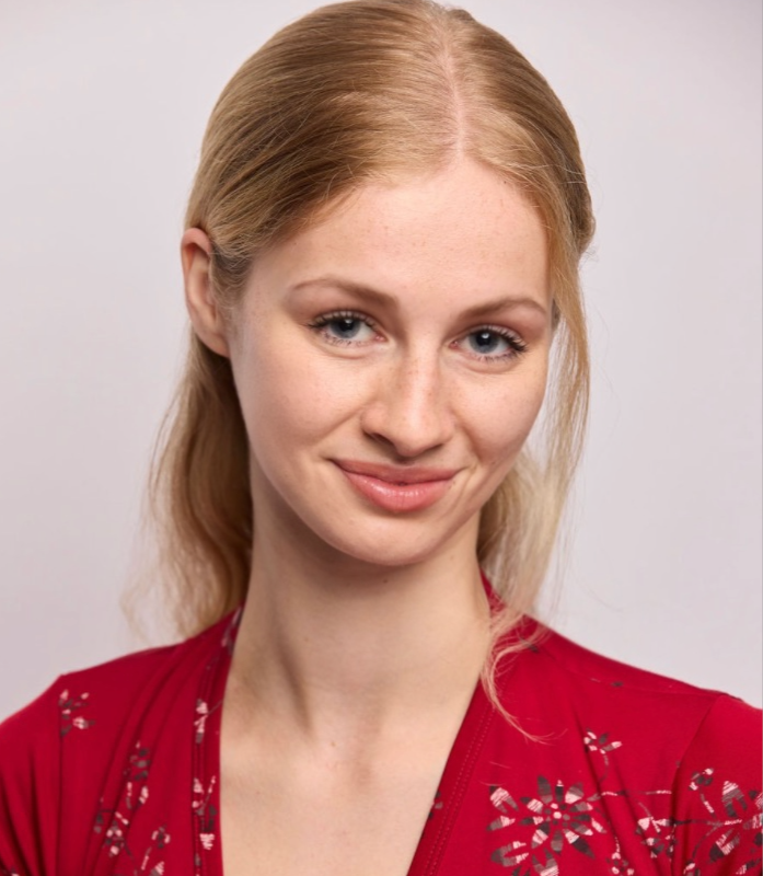 Annie Thomas, a young actress with light skin and blonde hair smiling, wearing a red top with floral details against a plain light background.
