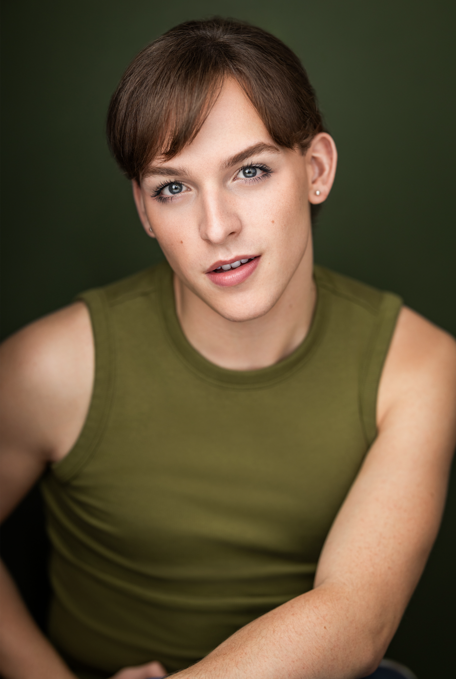 Gustavo Molina, a young actor with short brown hair and blue eyes, wearing a sleeveless green top, looking at the camera with a slight smile against a dark green background.