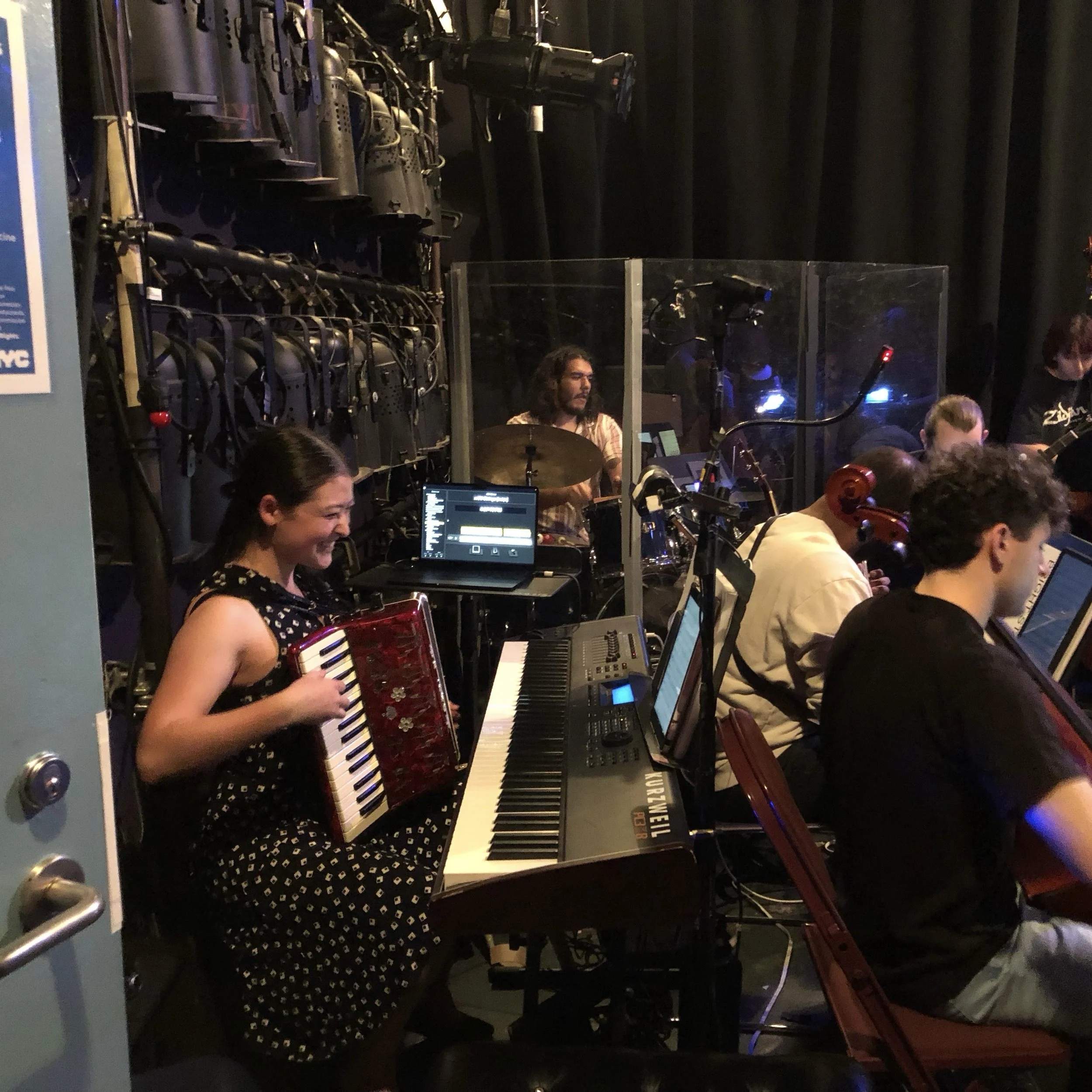 Sarah Chen playing a red accordion with a black and white keyboard in a band, with music equipment and other musicians in the background at a live music event.