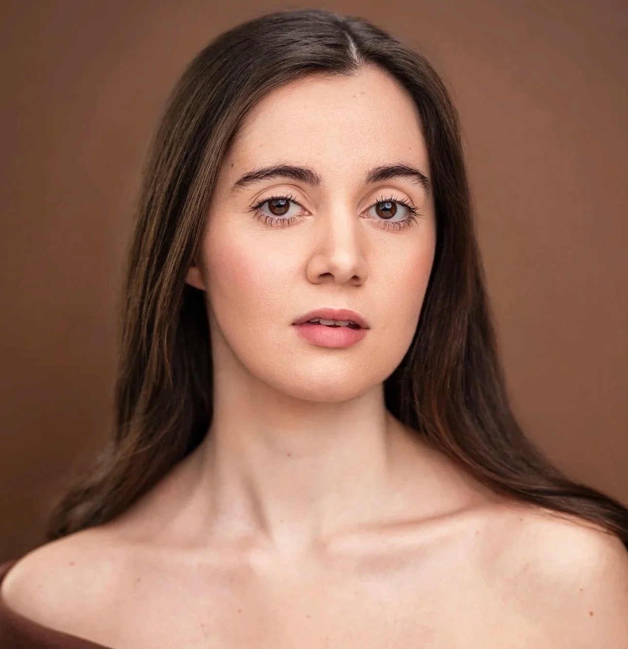 Close-up of Brooke Cassatto, a young actress with long brown hair and fair skin, looking directly at the camera with a neutral expression, against a plain brown background.