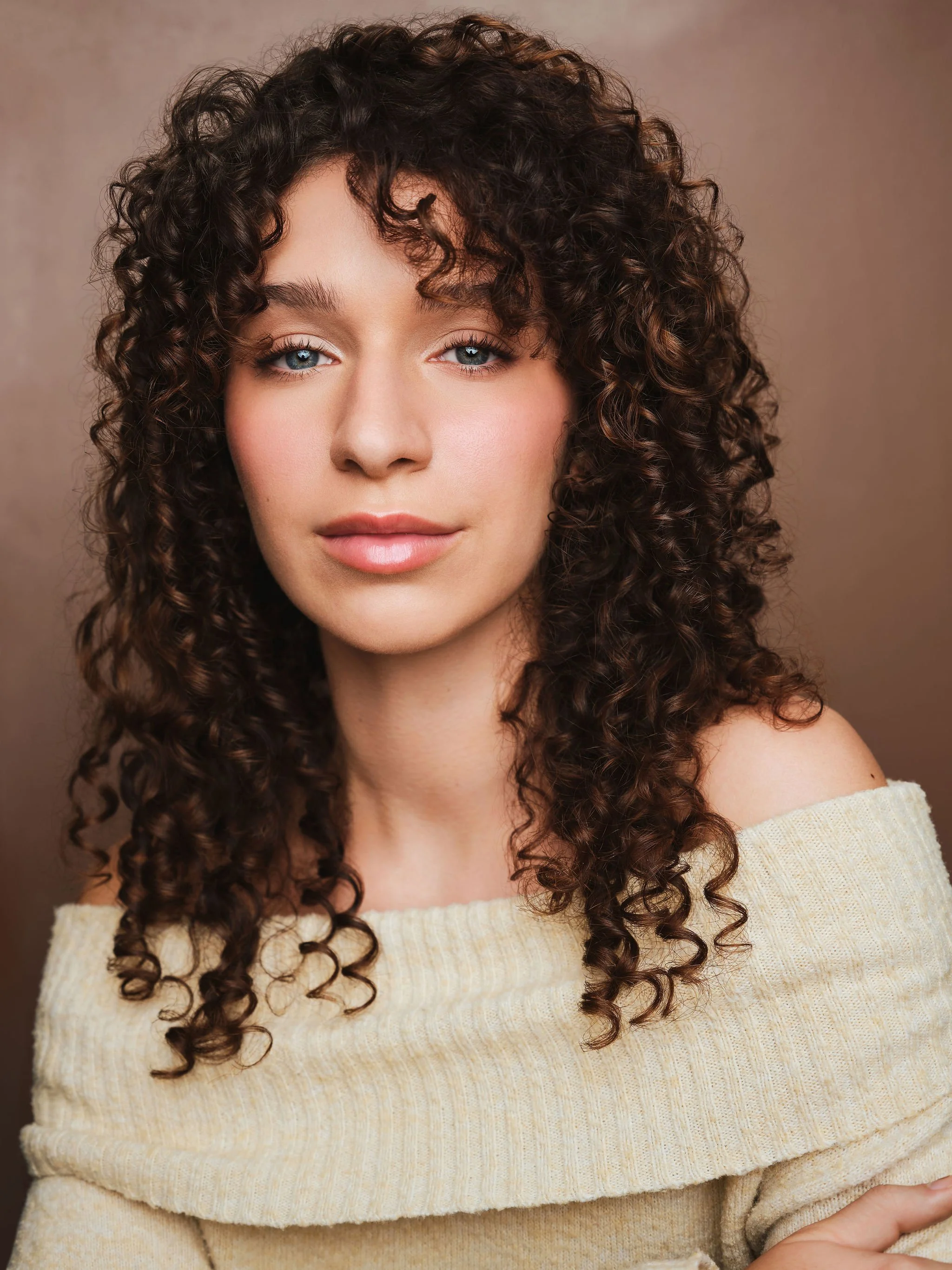 Rachel Rudolph, a young actress with curly brown hair and blue eyes, poses calmly against a brown backdrop wearing a tan sweater