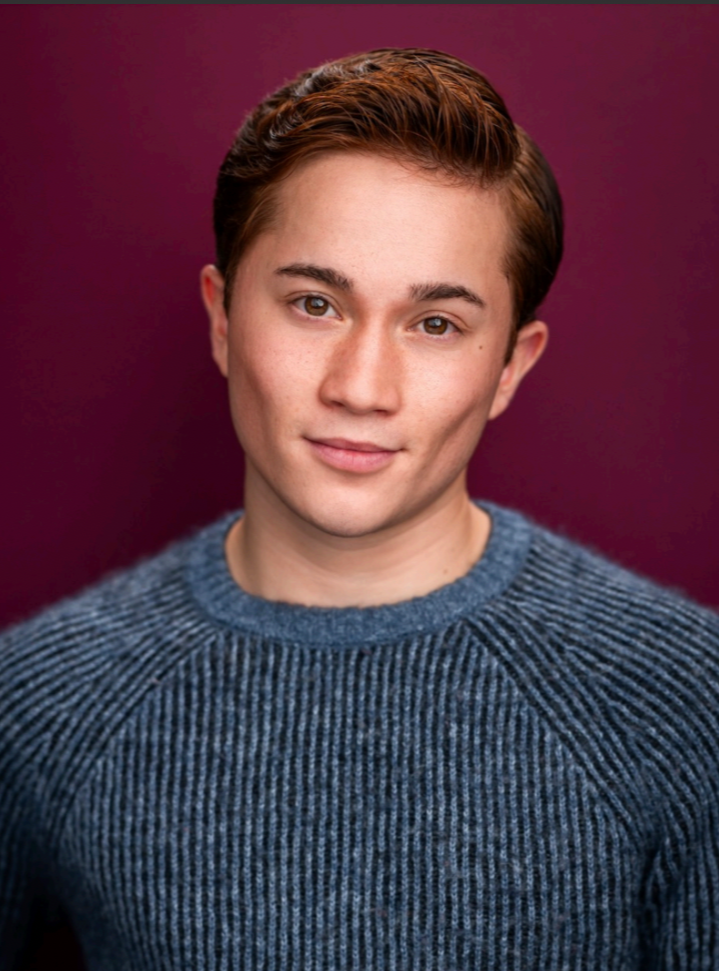 Noah Ong Bamola, a young actor with ginger hair and brown eyes poses calmly against a maroon backdrop with a blue sweater on