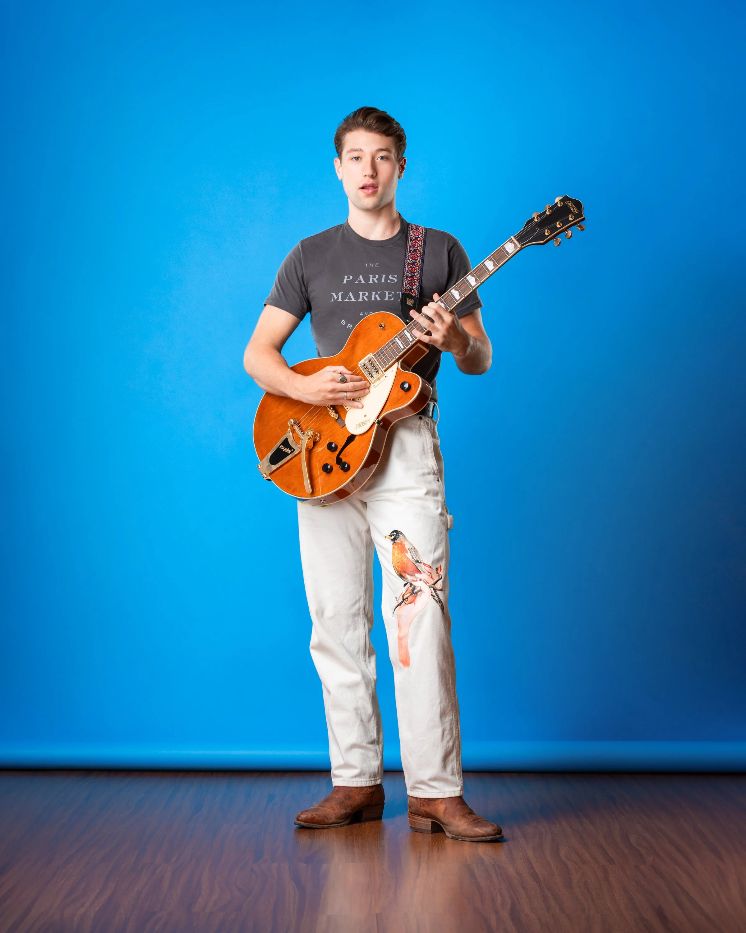 Jack Plogger, a young actor and musician, holding a guitar while standing against a bright blue backdrop wearing white jeans, brown boots, and a gray tee shirt