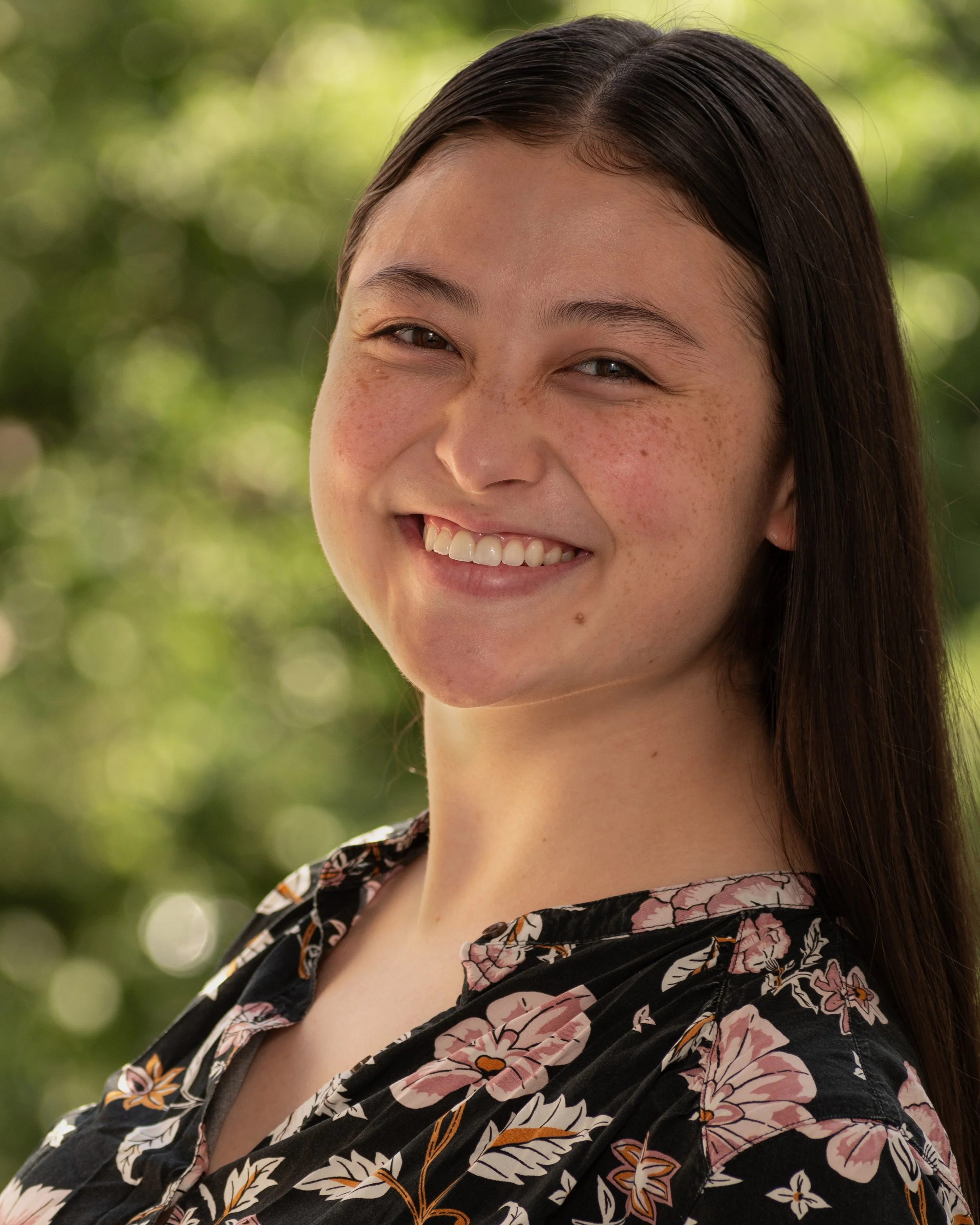 Sarah Chen, a young actress with dark brown hair and dark brown eyes smiling in a headshot, wearing a black top with a floral print, posed against a nature background