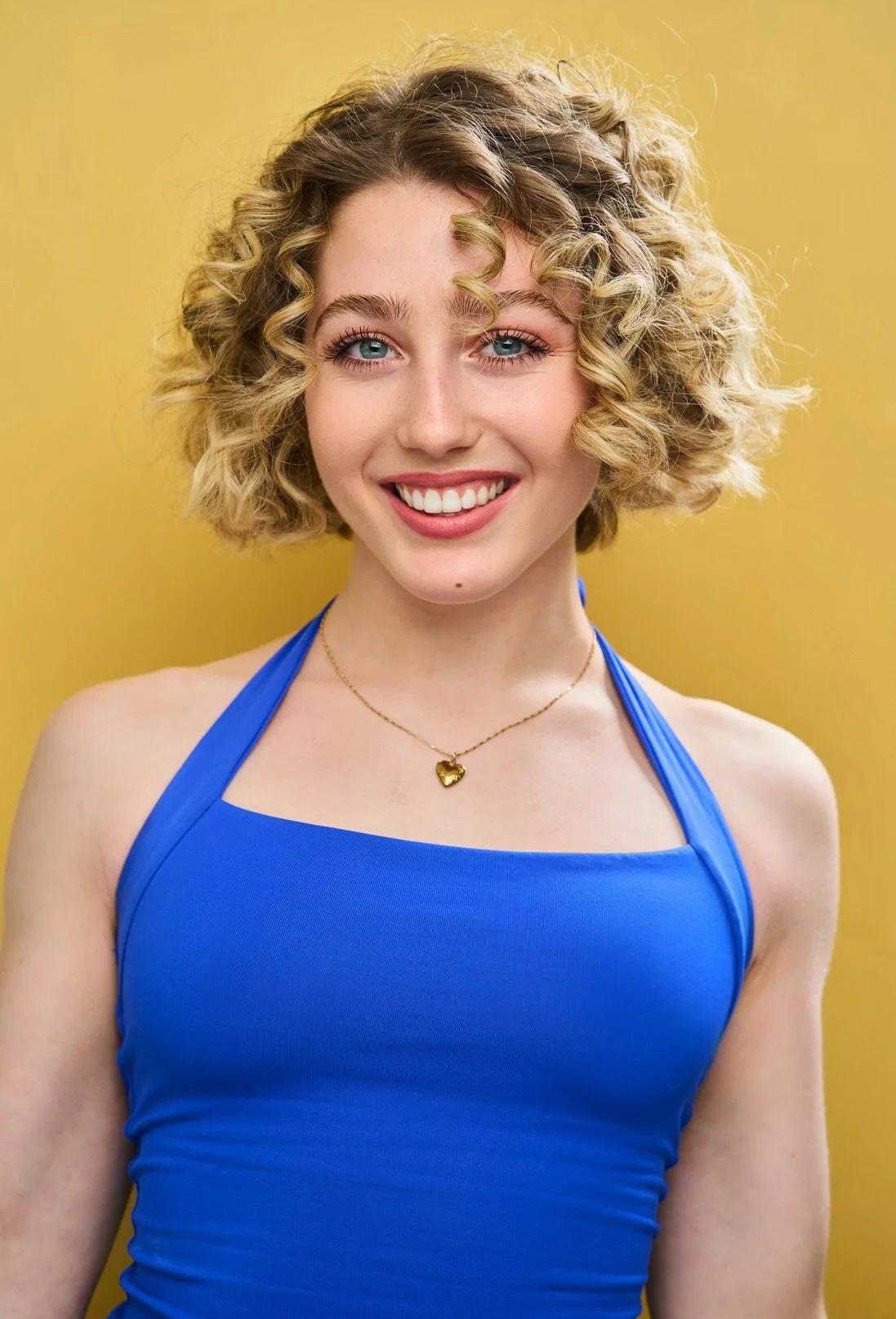 Brynn Ayoob, a young actress with curly blonde hair, blue eyes, and a bright smile wearing a blue tank top and a gold necklace with a heart pendant, standing against a yellow background.