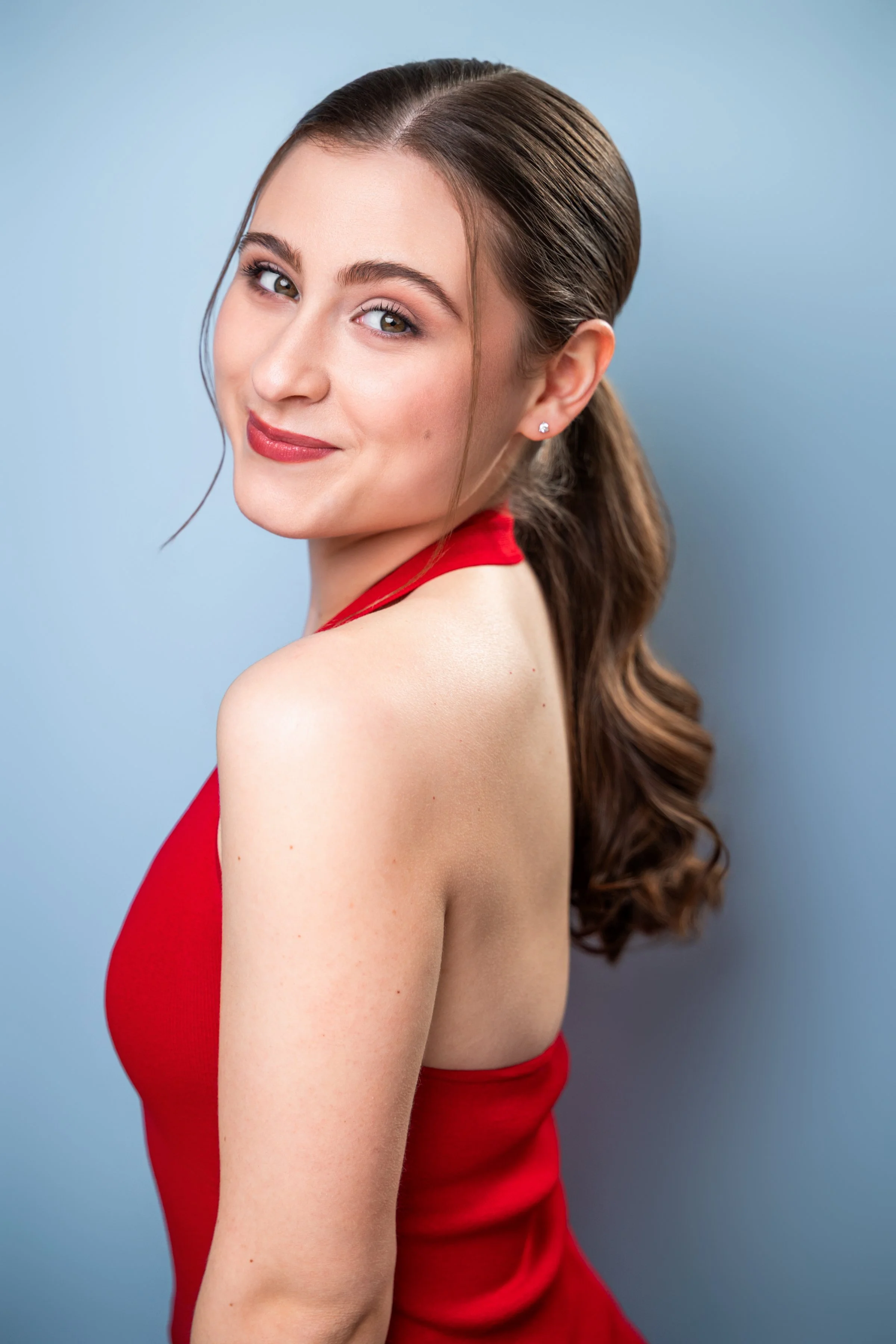 Maria Sablich, a young actress with a curled brown ponytail and brown eyes, in a red halter top against a light blue backdrop