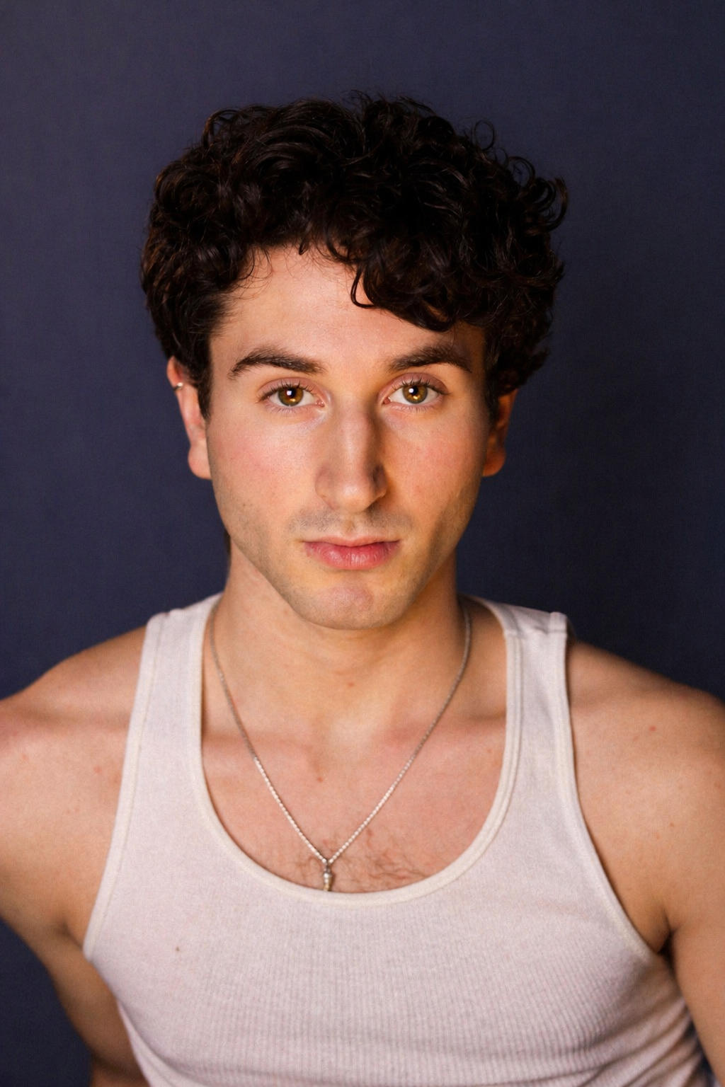 JJ Razzano, a young actor with curly dark hair, wearing a white tank top and a silver necklace, smiling slightly against a dark background.