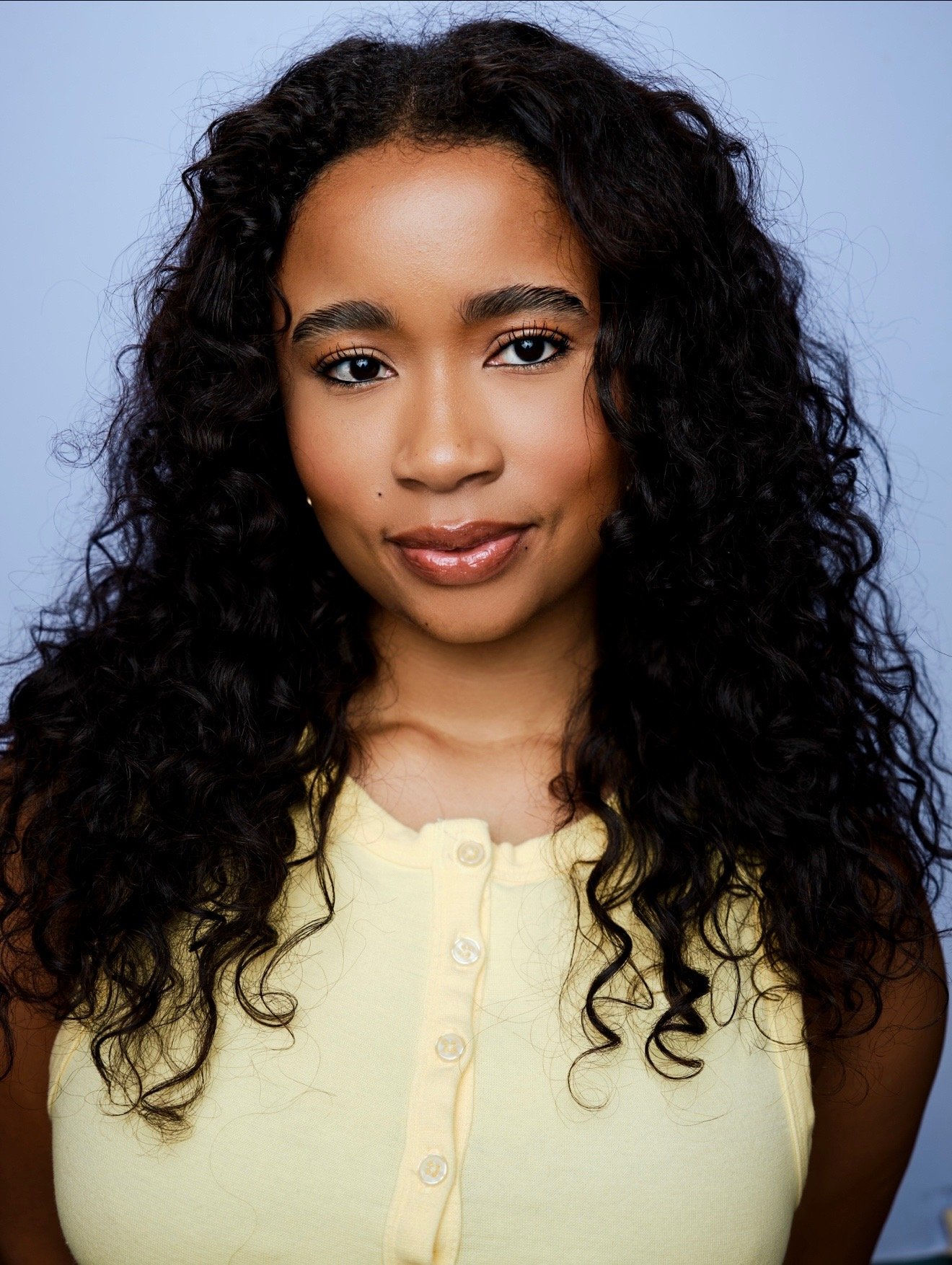 Tuesday Usry, a young actress with dark curly hair and brown eyes smiling in a butter yellow top against a light blue backdrop