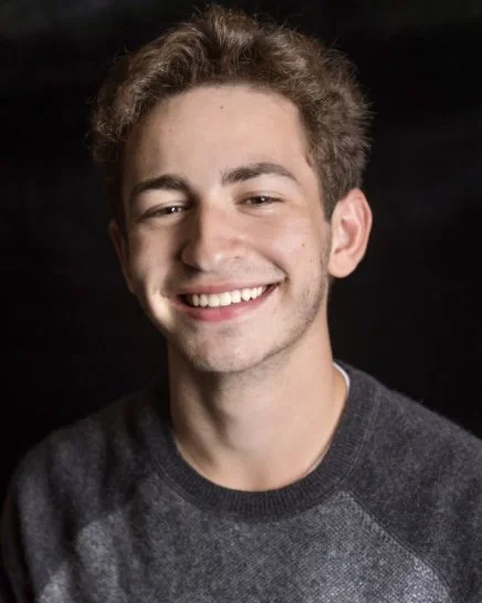 Davey Fried, a young actor with brown hair and brown eyes in a gray top against a black backdrop
