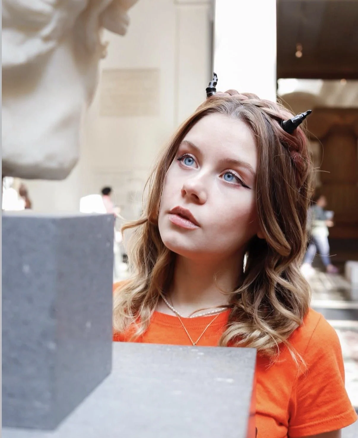 Lizzie Matlock, a young actress with wavy blonde hair and blue eyes, wearing an orange shirt, looks at a stone sculpture of a head in a museum or gallery.