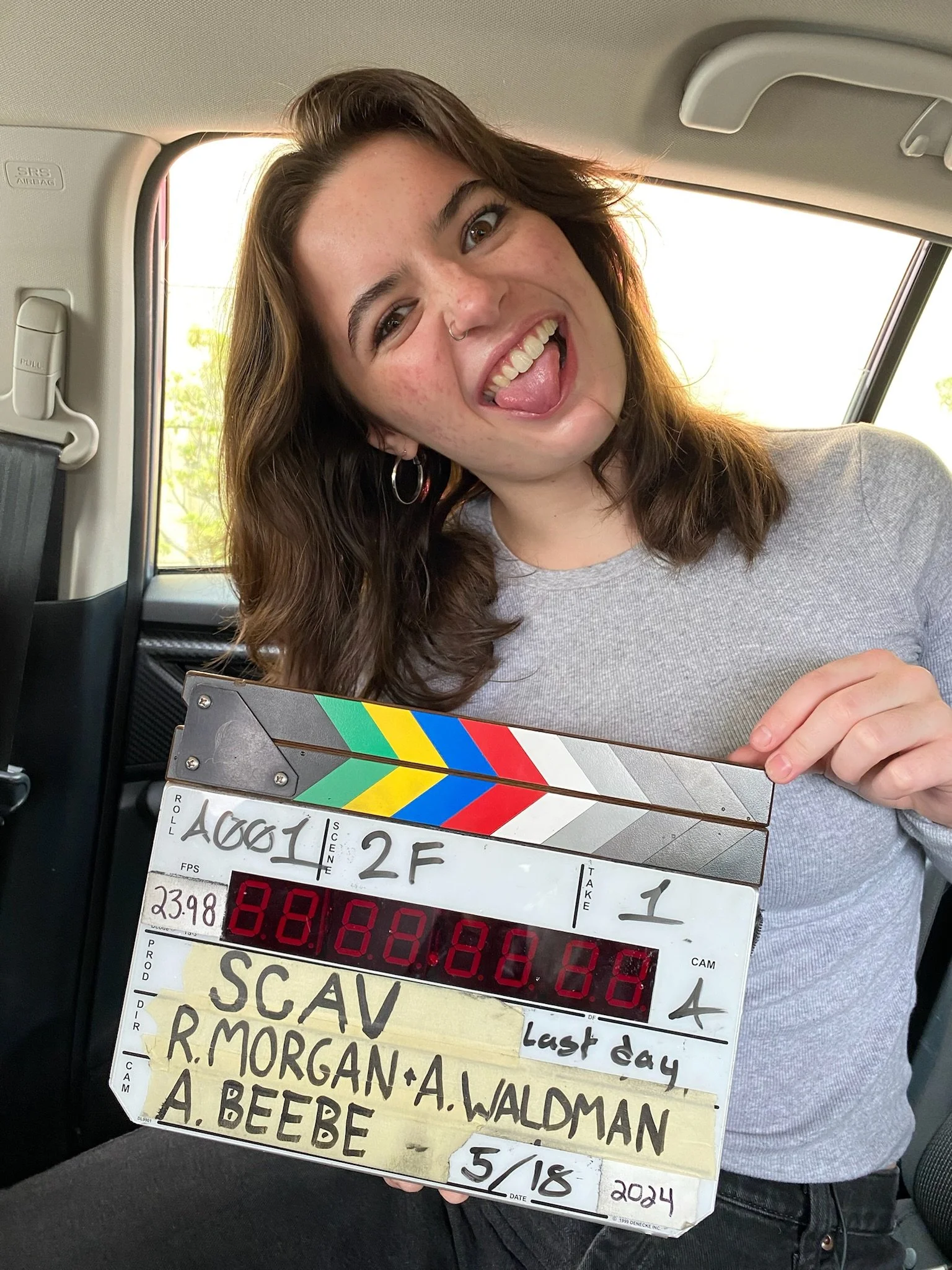 Kelsey LeVie, a young actress, holding a director's clapboard on a car seat, smiling and sticking her tongue out.