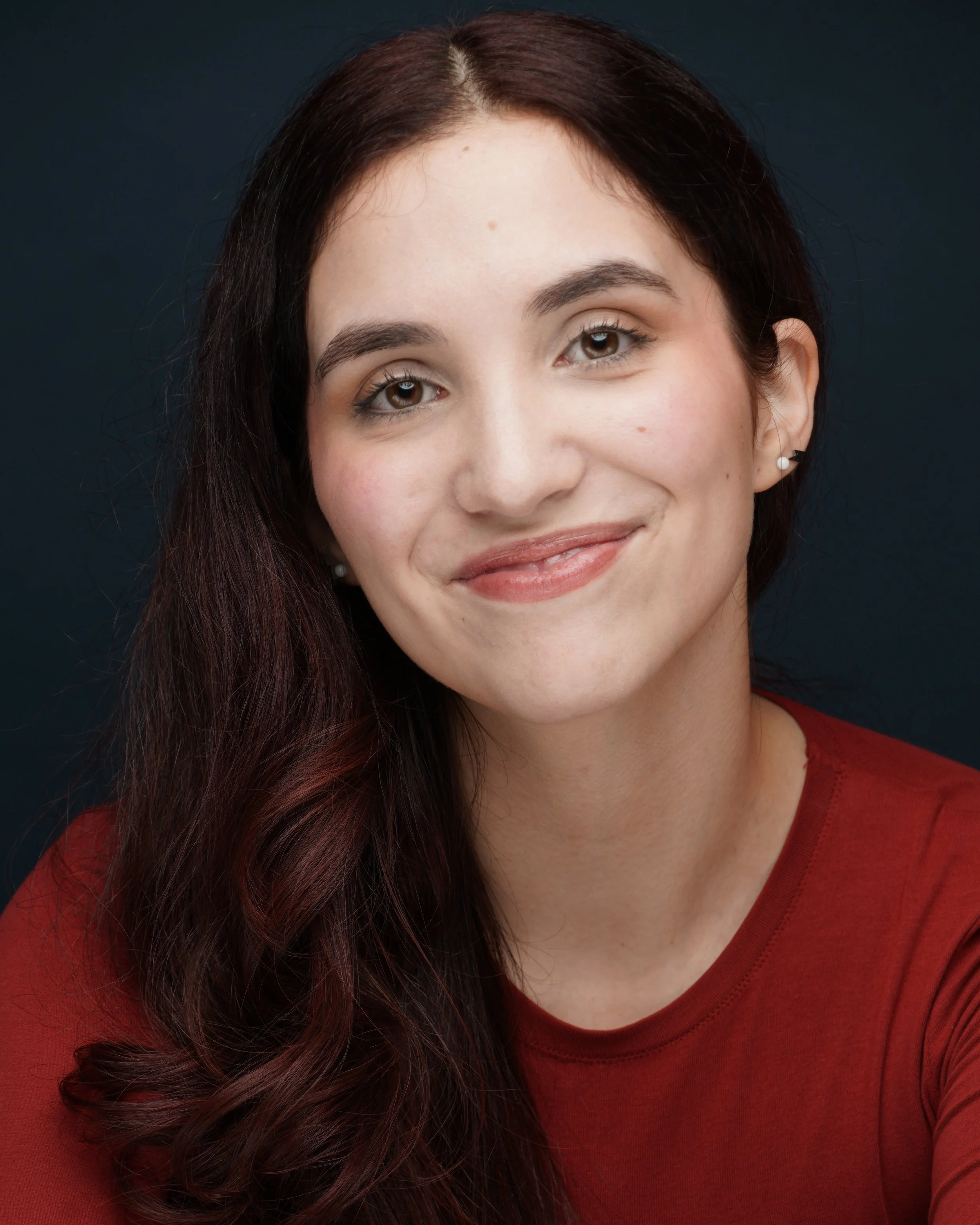 Close-up of Andrea Mongil, a young actress with dark red hair, light skin, wearing pearl earrings and a red shirt, smiling, against a dark background.