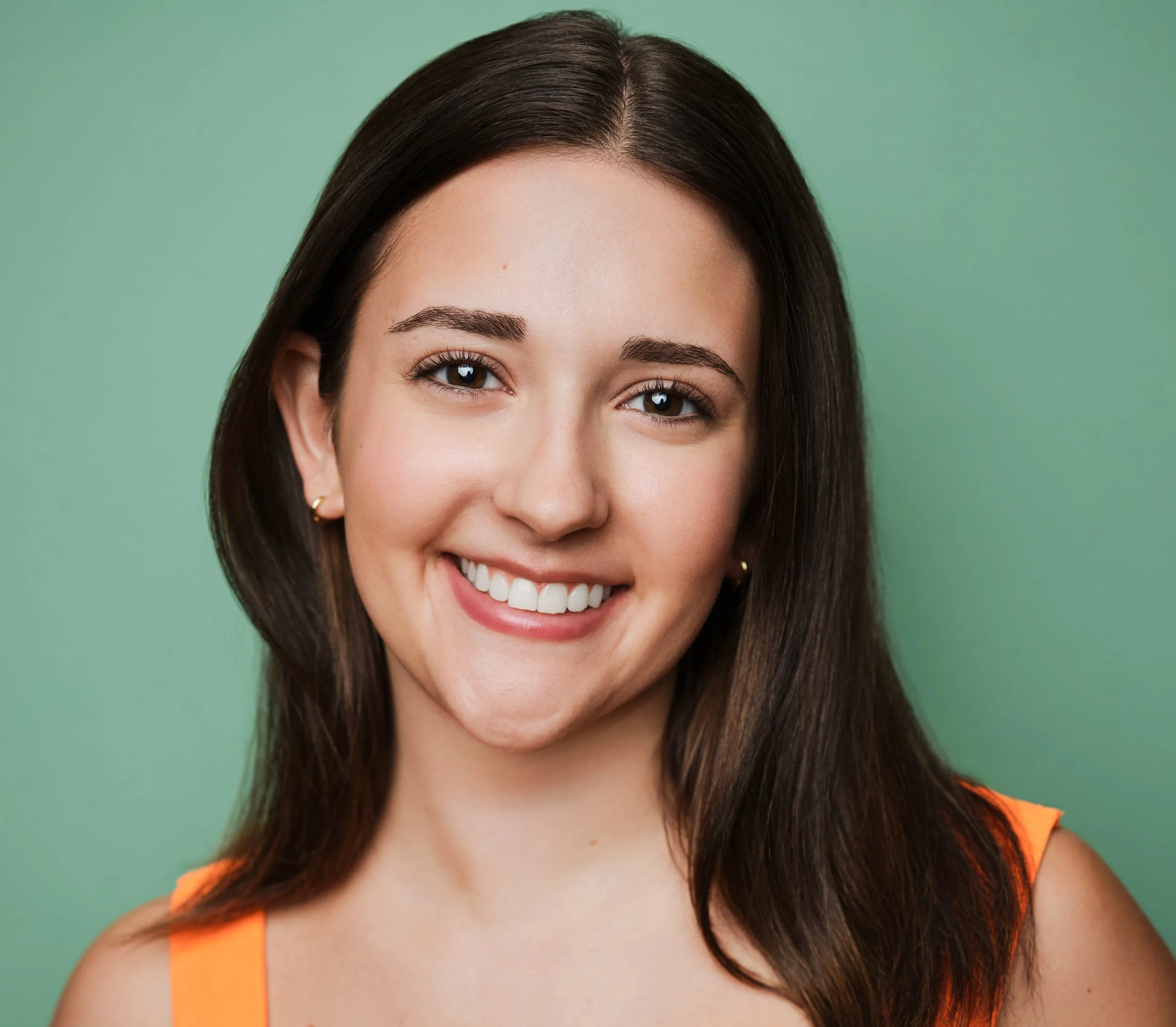 Close-up of Jessica Schuchardt, a young actress with long dark hair, wearing a bright orange top, smiling against a green background.