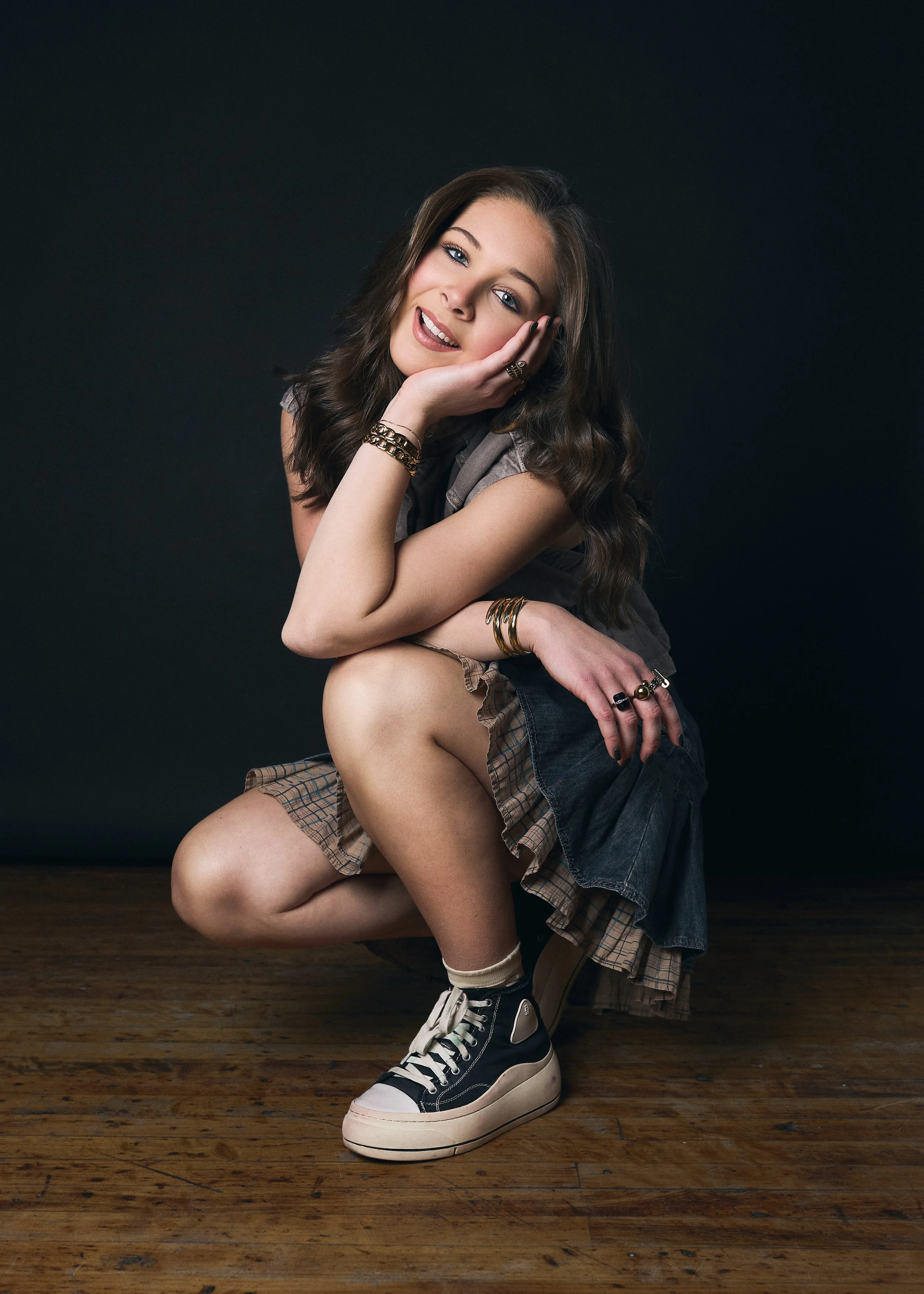 Lucy McIver, a young actress with brown hair and blue eyes, poses in a squat while leaning on her hand, wearing a denim and plaid layered skirt, Converse sneakers, and bracelets and rings