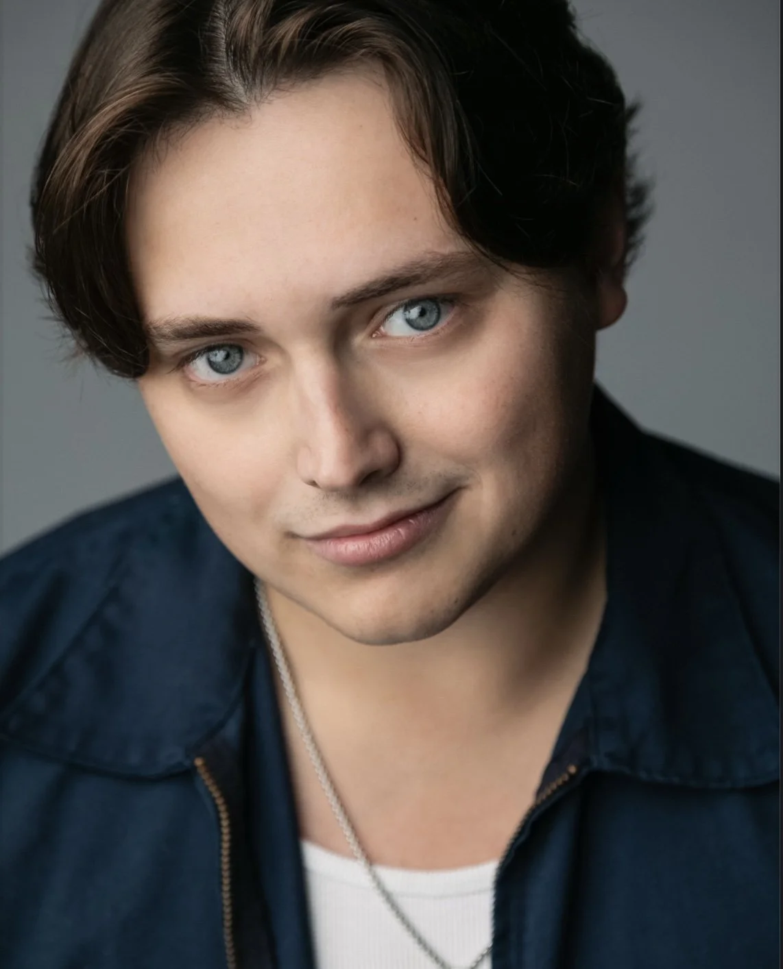Brandon Berg, a young actor with brown hair and blue eyes, in a white top and navy jacket against a gray backdrop