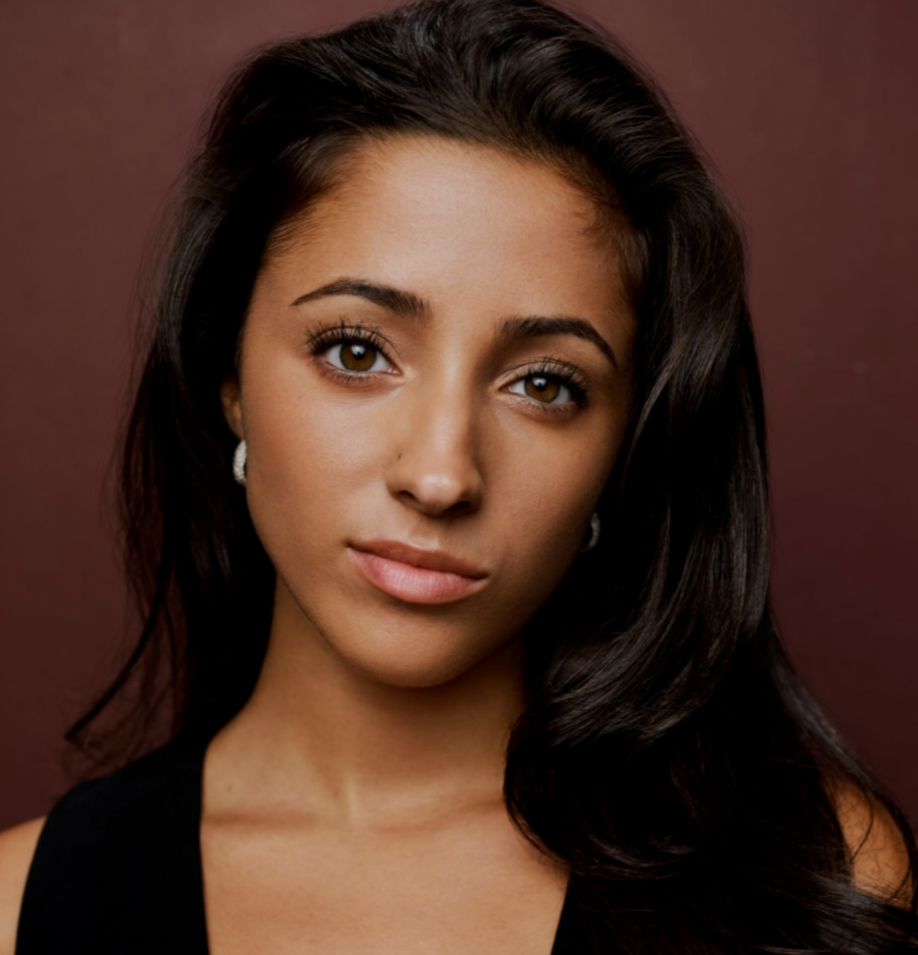 Close-up portrait of Elana Simard, a young actress with long dark hair, brown eyes, and light makeup, wearing earrings and a black top, in front of a maroon background.