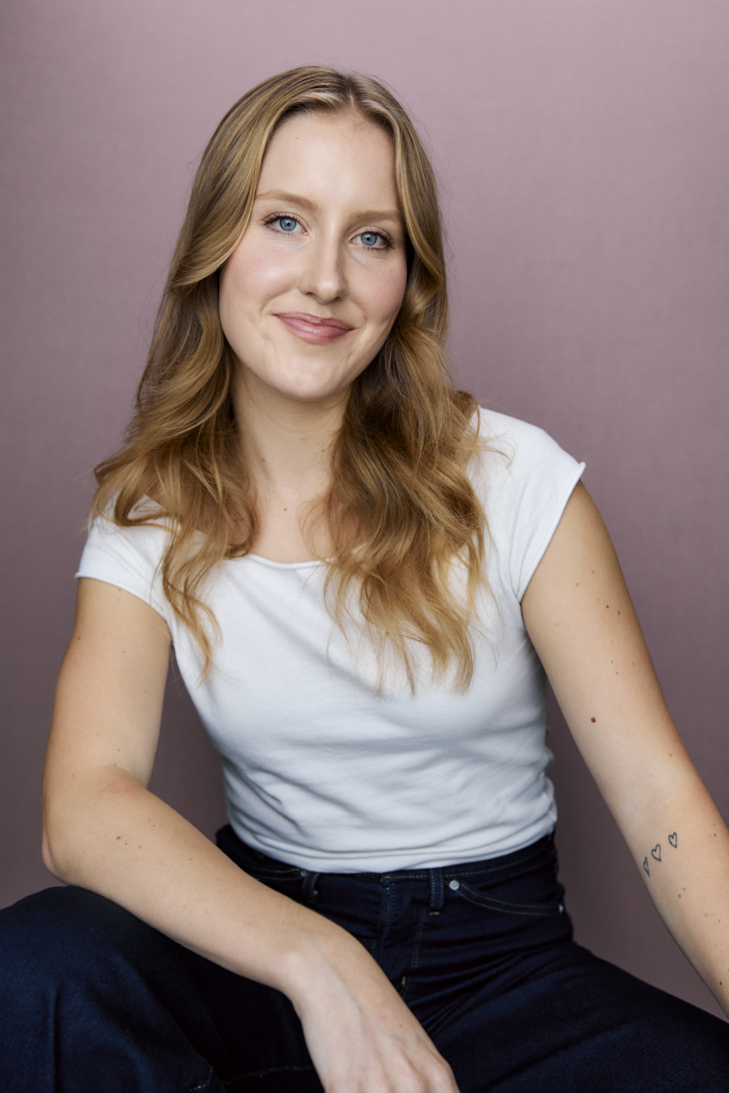 Mimi Pietila, a young actress with wavy blonde hair and blue eyes, poses with a soft smile in a relaxed white top against a mauve backdrop